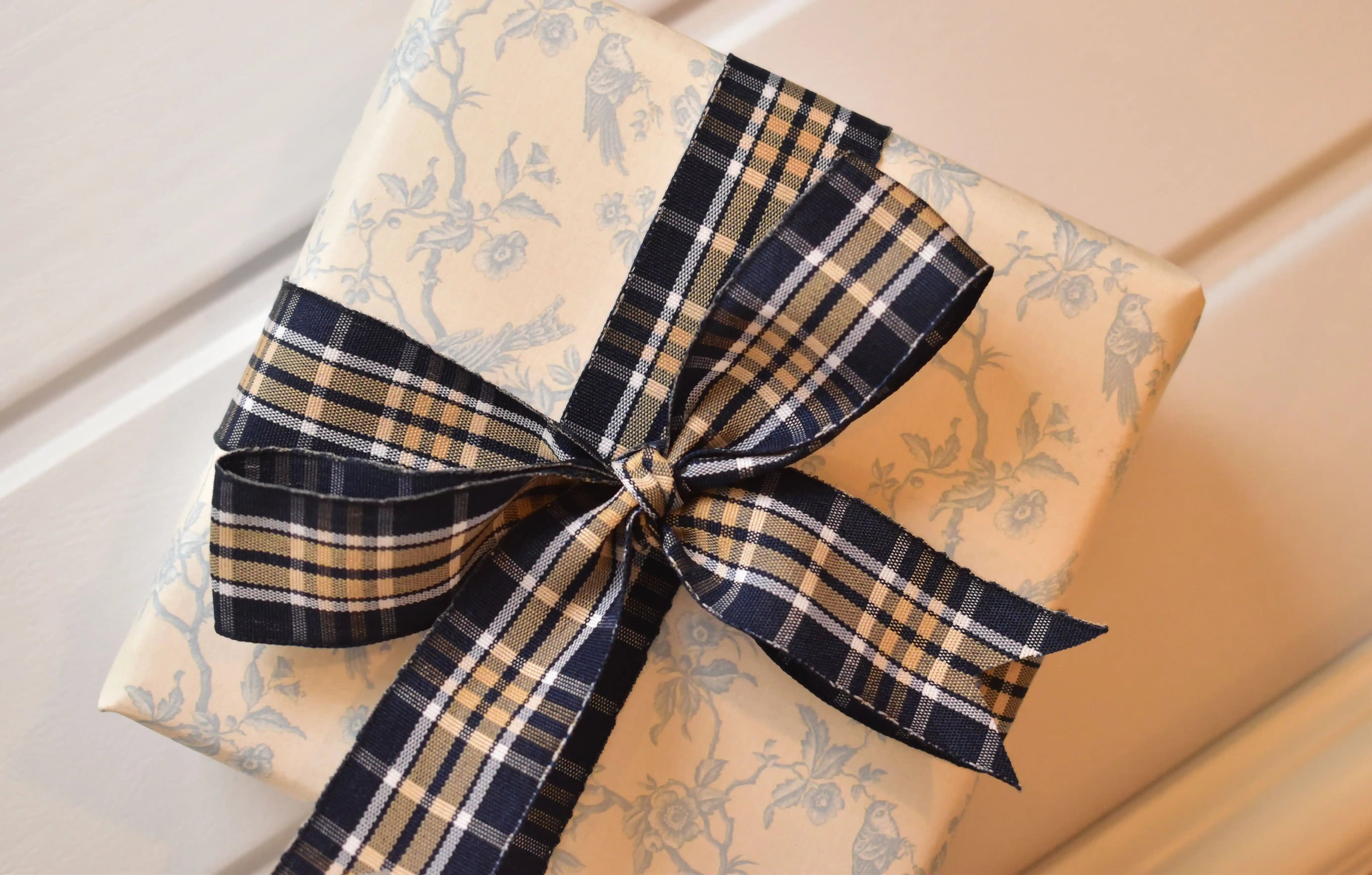 This image displays the Midnight Plaid ribbon, showcasing its rich navy and white plaid design and woven texture. A long strand is loosely unravelled to highlight the full pattern, with a section wrapped around a "Sunlit Hugs" brand tag.