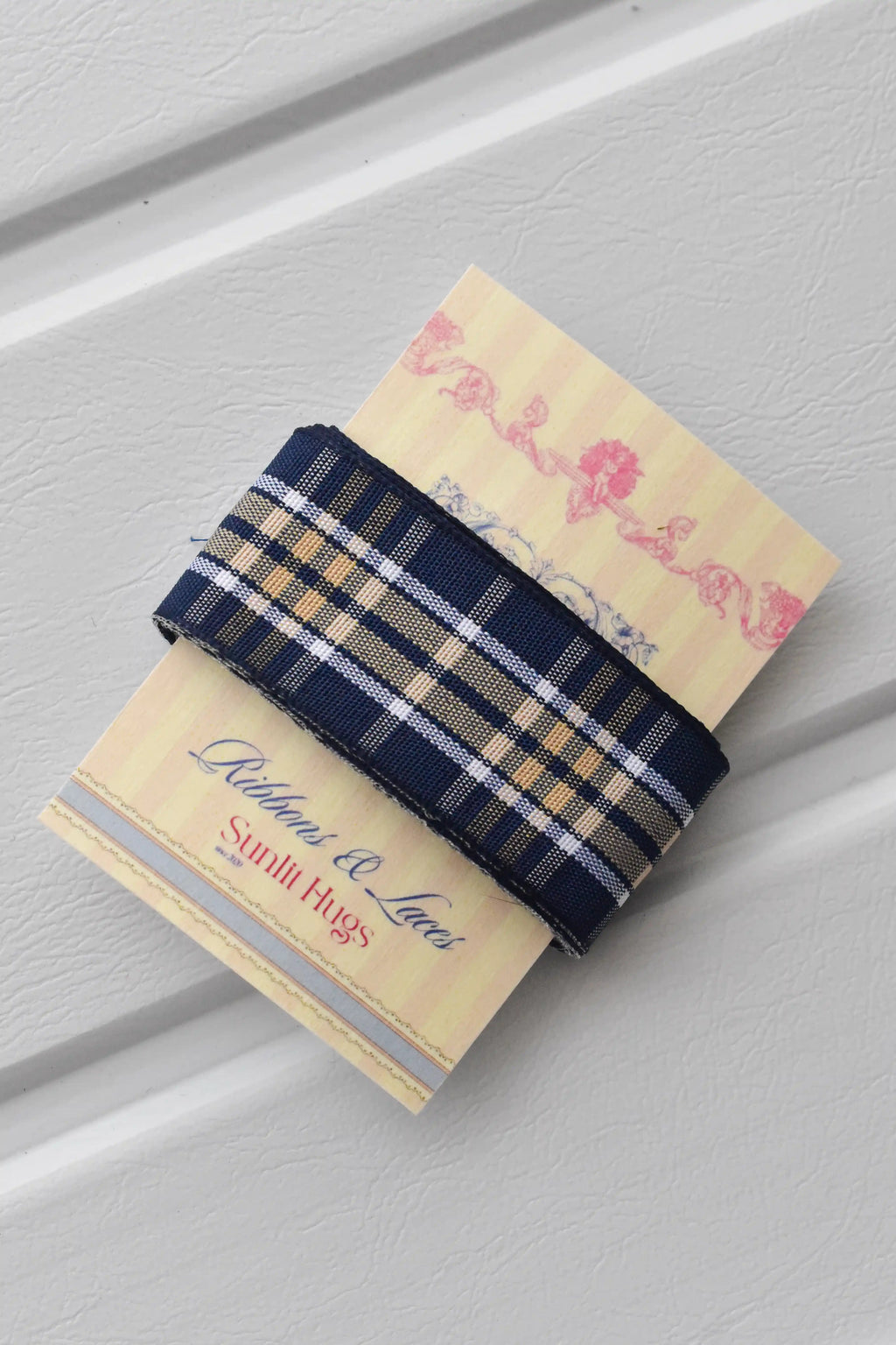 This detailed, head-on view of the Midnight Plaid ribbon highlights its refined navy and white plaid pattern. The ribbon is wrapped tidily around the "Sunlit Hugs" brand tag to show its precise design and colours.