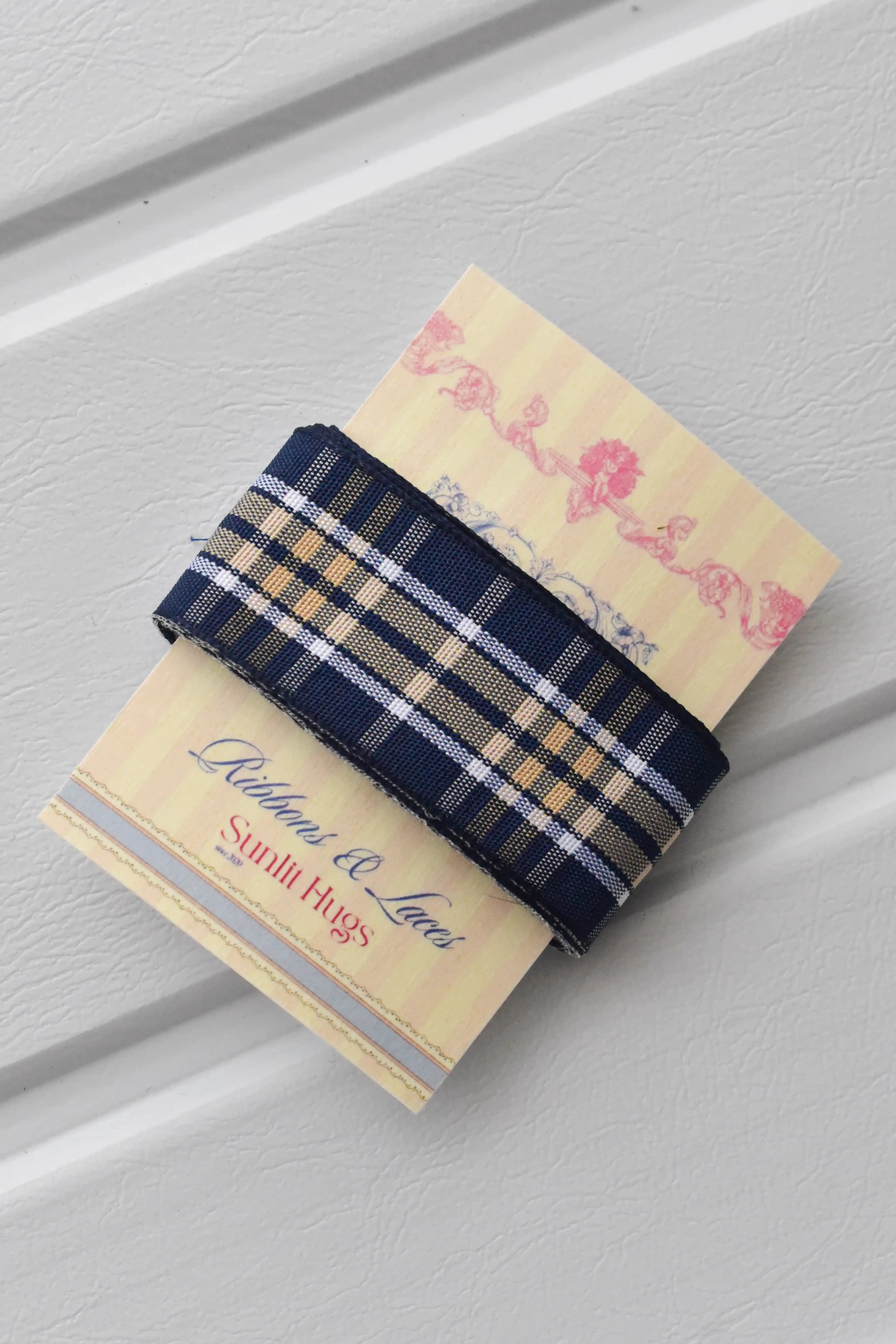 This detailed, head-on view of the Midnight Plaid ribbon highlights its refined navy and white plaid pattern. The ribbon is wrapped tidily around the "Sunlit Hugs" brand tag to show its precise design and colours.