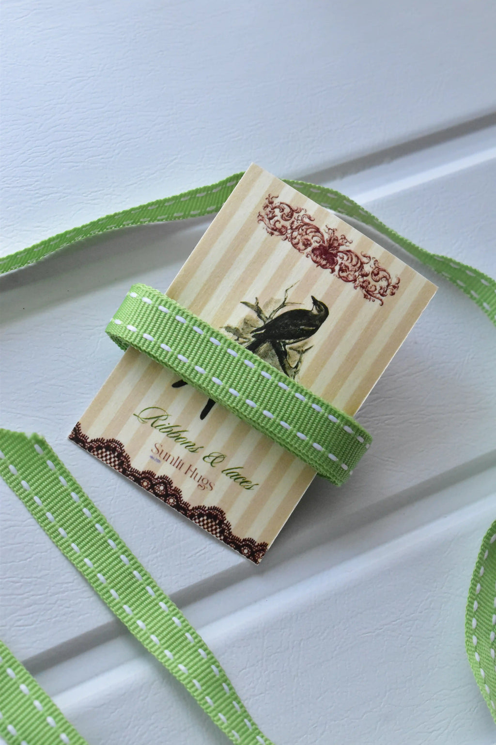 This image displays the Moss Stitch ribbon, showcasing its beautiful pastel green colour and delicate white dashed stitching. A long strand is loosely unravelled to highlight the full design, with a section wrapped around a "Sunlit Hugs" brand tag.