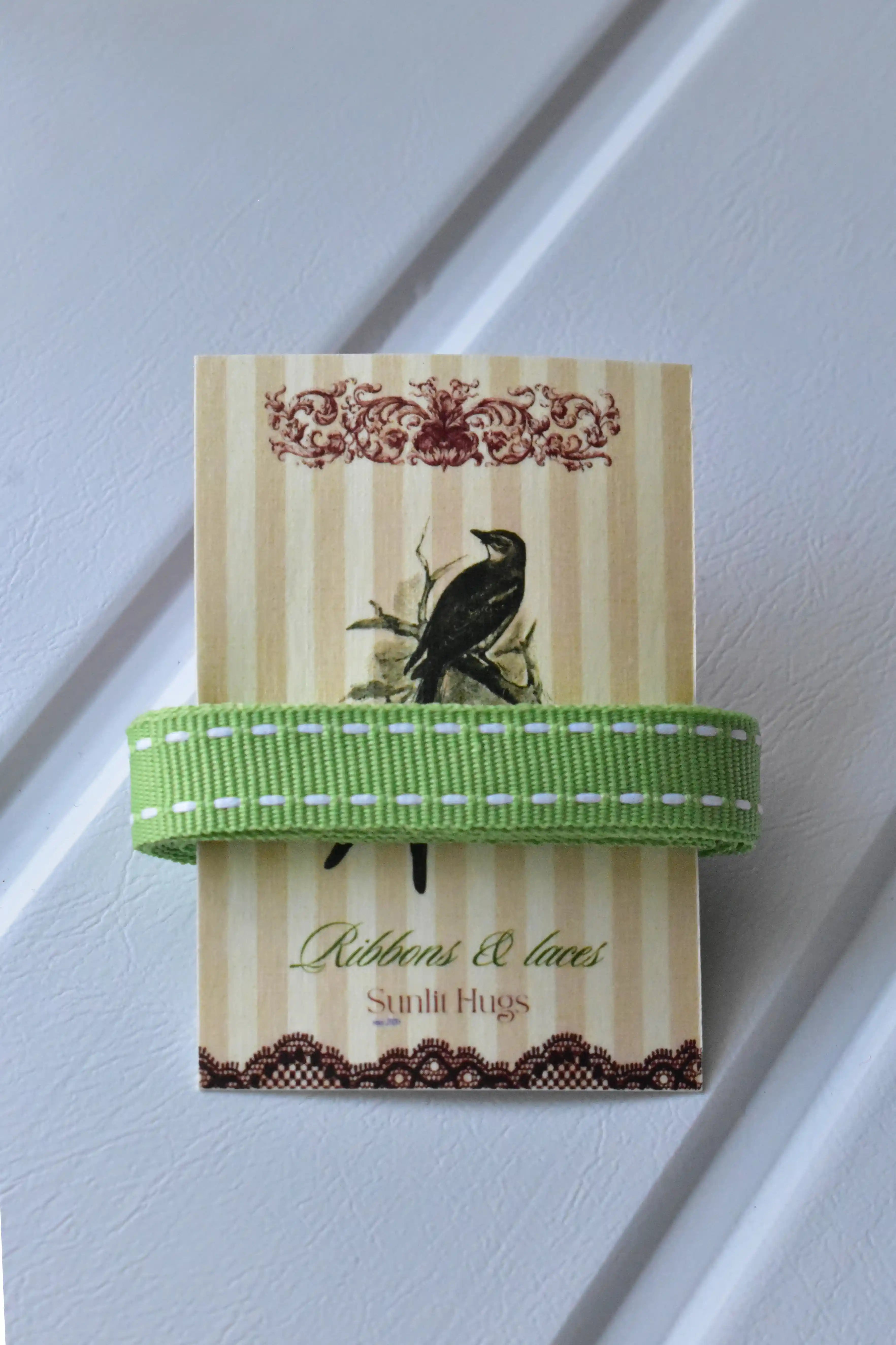 This detailed, head-on view of the Moss Stitch ribbon highlights its soft pastel green colour and the contrasting white dashed stitching. The ribbon is wrapped tidily around the "Sunlit Hugs" brand tag to show its precise design.