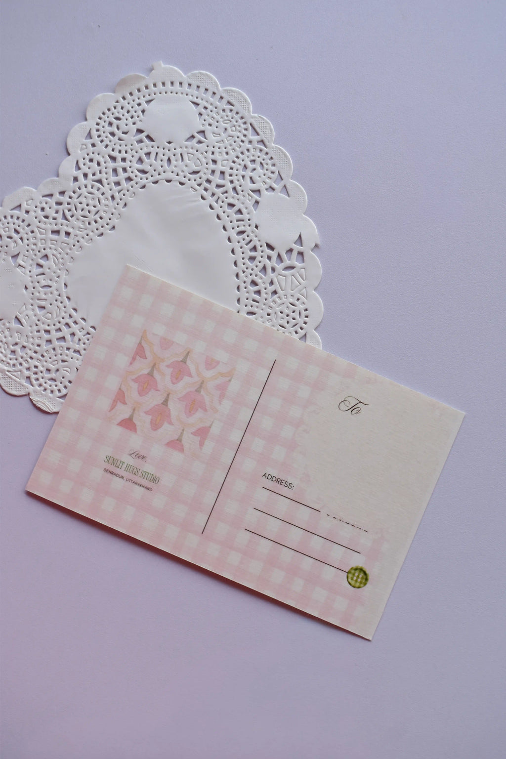 The back view of a postcard resting on a white paper doily. The design is a soft pink and white wide gingham pattern, with a central square featuring a pink floral bell pattern. It includes labeled fields for address and postage.