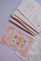 A stack of postcards showing the pink gingham reverse design on the top cards, with one card laid flat displaying the 'Find beauty in mundane' vintage pattern front. The cards have a wide pink and white check pattern.