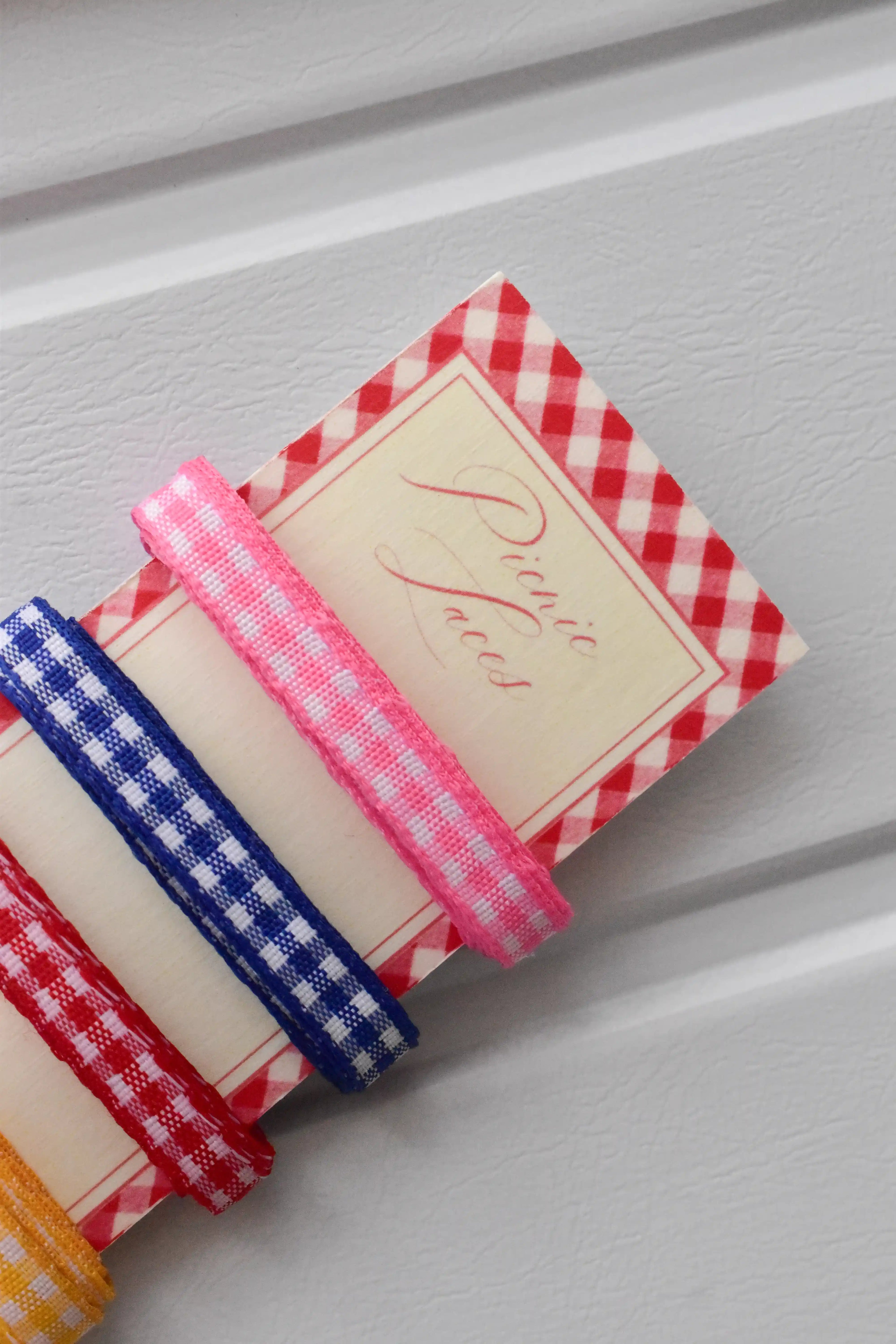 Angled close-up of the pink, blue, and red narrow gingham ribbons wrapped around the top corner of the "Picnic Laces" display card.