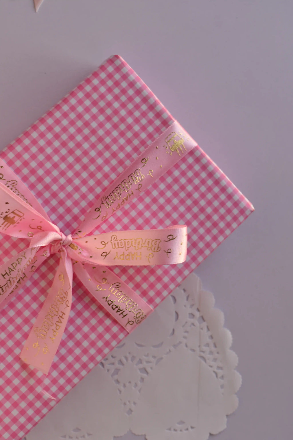 Close-up of a pink checkered gift box wrapped with the soft pink birthday ribbon featuring gold foil script and tied in a beautiful bow.