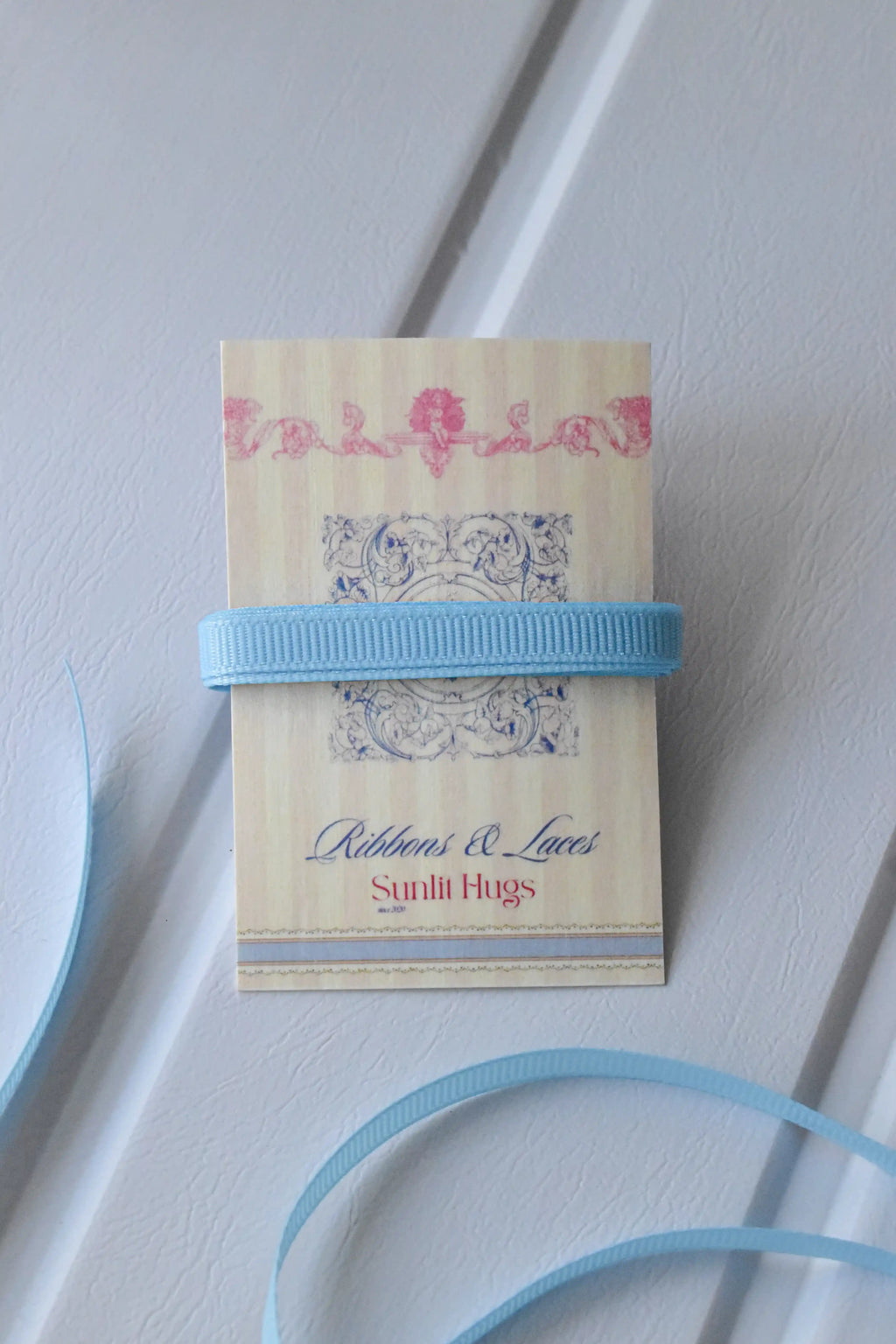 This image displays the Powder Blue Charm ribbon, showcasing its beautiful light blue colour and delicate texture. A long strand is loosely unravelled to highlight the full design, with a section wrapped around a "Sunlit Hugs" brand tag.