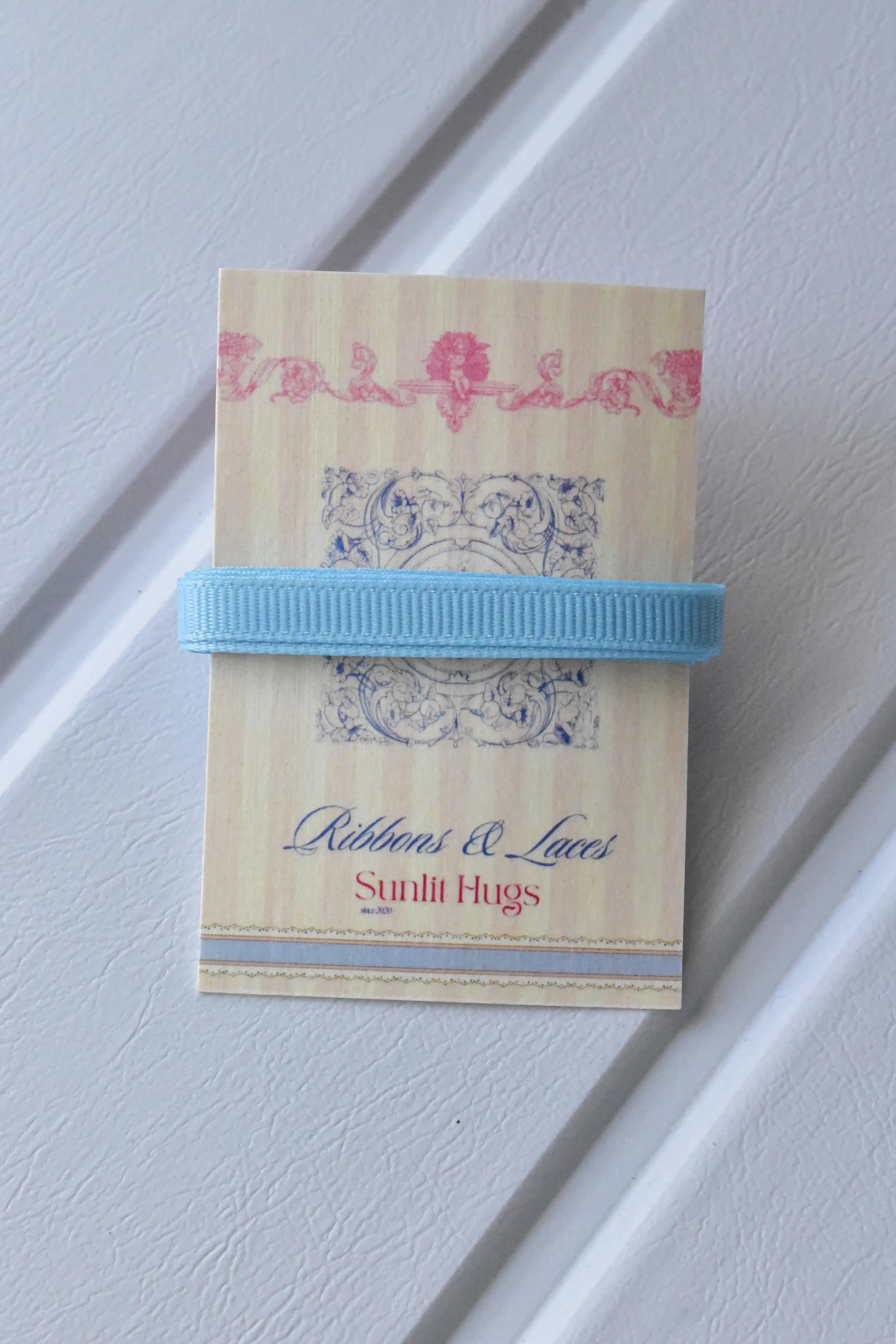 This detailed, head-on view of the Powder Blue Charm ribbon highlights its soft light blue colour and grosgrain texture. The ribbon is wrapped tidily around the "Sunlit Hugs" brand tag to show its precise design.
