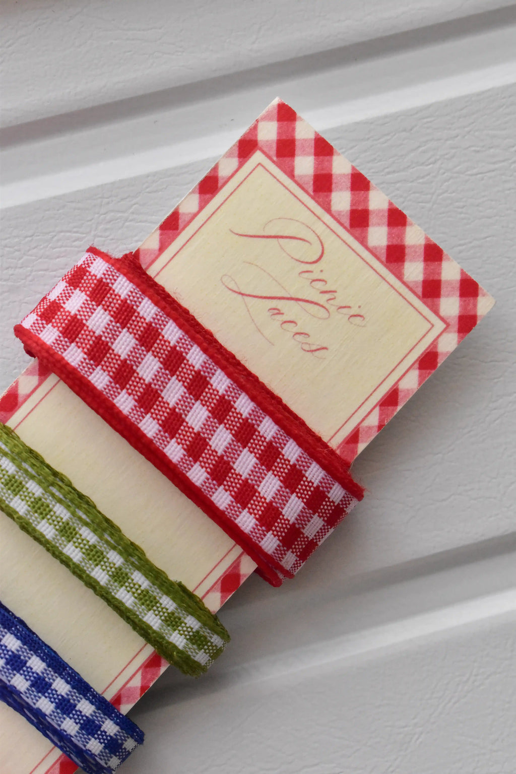 Angled close-up of the red and green narrow gingham ribbons wrapped around the top corner of the "Picnic Laces" display card.