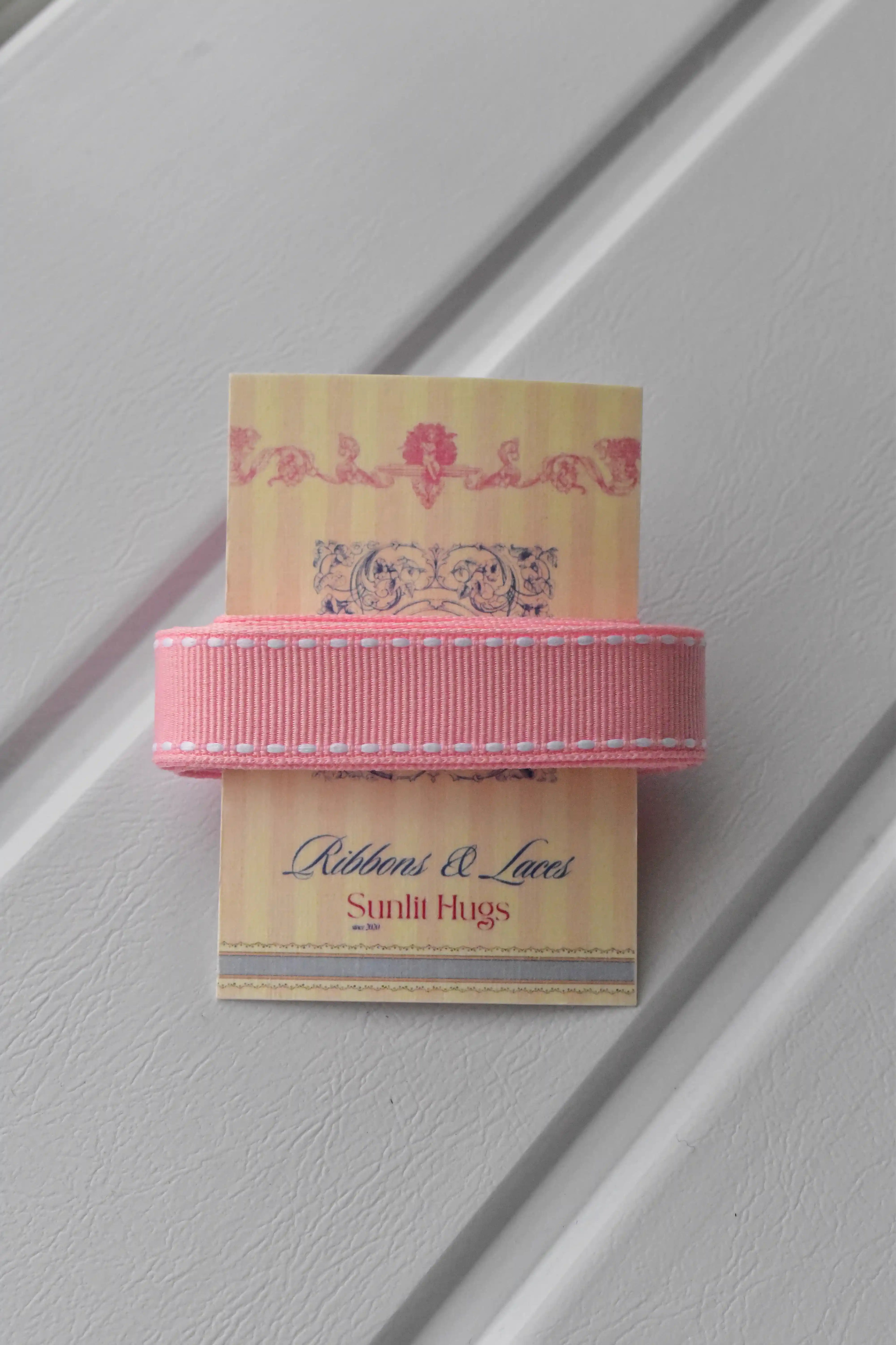 This detailed, head-on view of the Rose Taffy ribbon highlights its soft pastel pink colour and the contrasting white dashed stitching. The ribbon is wrapped tidily around the "Sunlit Hugs" brand tag to show its precise design.