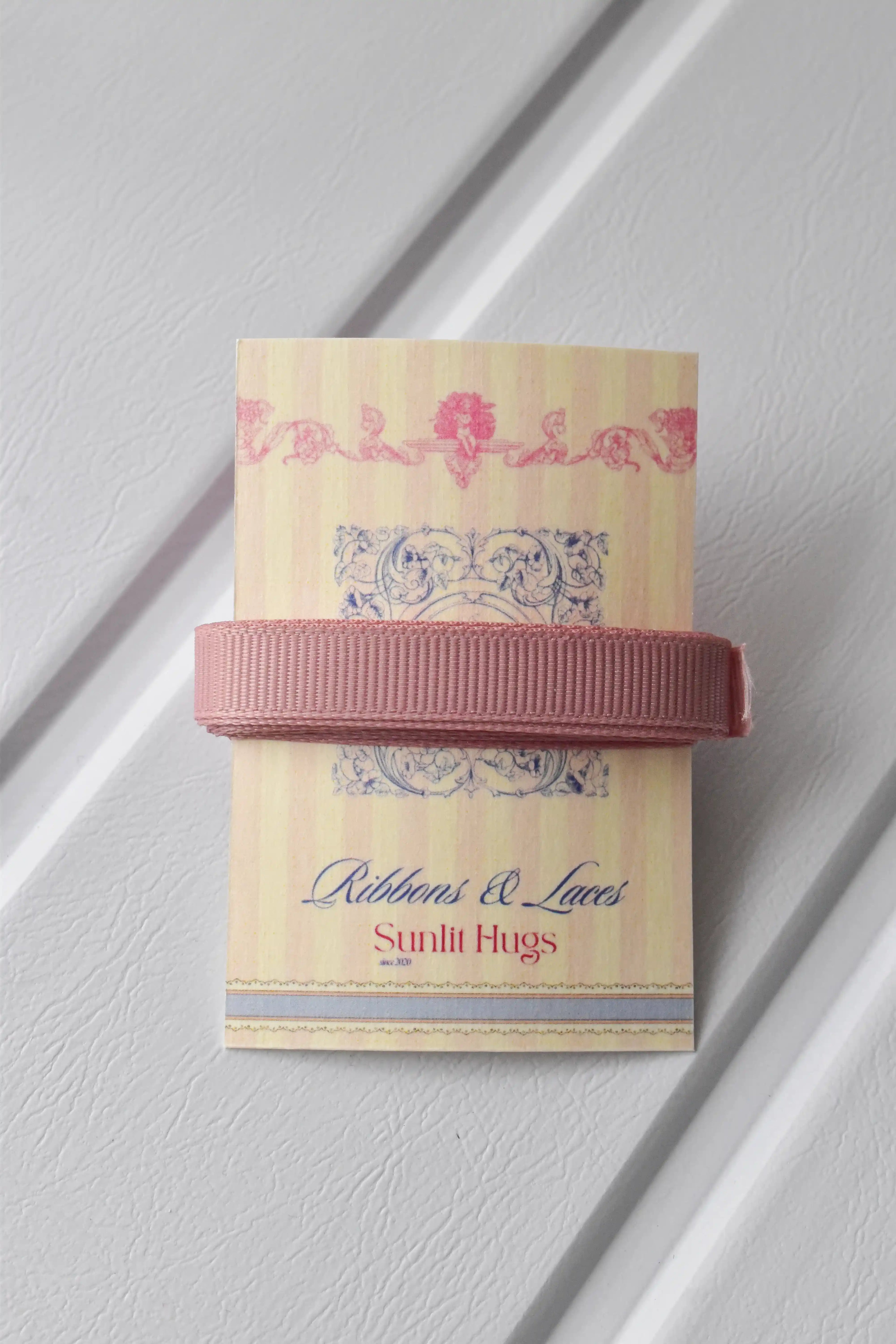 This detailed, head-on view of the Rustic Blush ribbon highlights its sophisticated rose gold colour and subtle texture. The ribbon is wrapped tidily around the "Sunlit Hugs" brand tag to show its precise design.