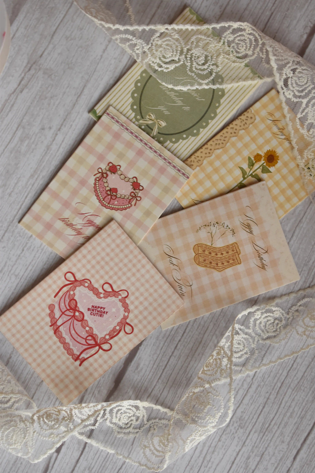 Flat lay of a set of 5 Sunlit Hugs Happy Birthday cards featuring vintage gingham and striped designs with illustrations of a ribbon heart, strawberry cake, vintage socks, and a sunflower, arranged with white floral lace on a wooden surface.