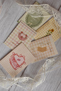 Flat lay of a set of 5 Sunlit Hugs Happy Birthday cards featuring vintage gingham and striped designs with illustrations of a ribbon heart, strawberry cake, vintage socks, and a sunflower, arranged with white floral lace on a wooden surface.