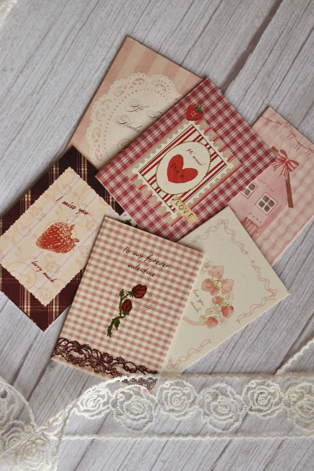 Flat lay of the Sunlit Hugs Set of 6 Love Series Cards featuring vintage romantic designs like strawberries, red plaid, pink gingham, and floral lace, arranged on a white wooden table with decorative lace.