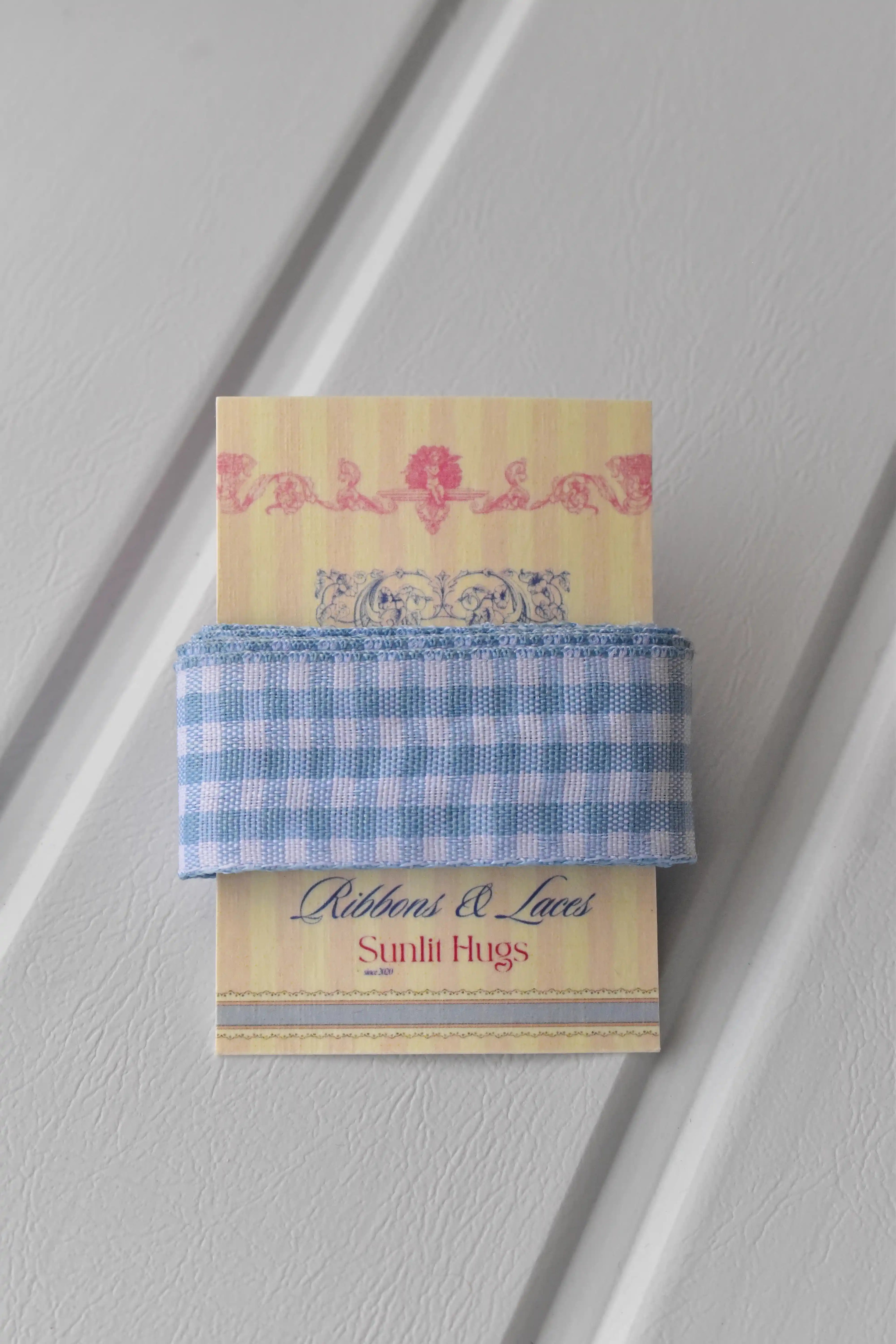 This detailed, head-on view of the Sky Gingham ribbon highlights its soft light blue and white checkered pattern. The ribbon is wrapped tidily around the "Sunlit Hugs" brand tag to show its precise design.
