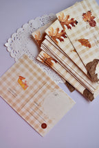 A stack of autumn postcards showing the front design of scattered brown and amber leaves and the emotional text. One postcard is turned over, displaying the pie slice and the address lines against the tan and cream gingham.

