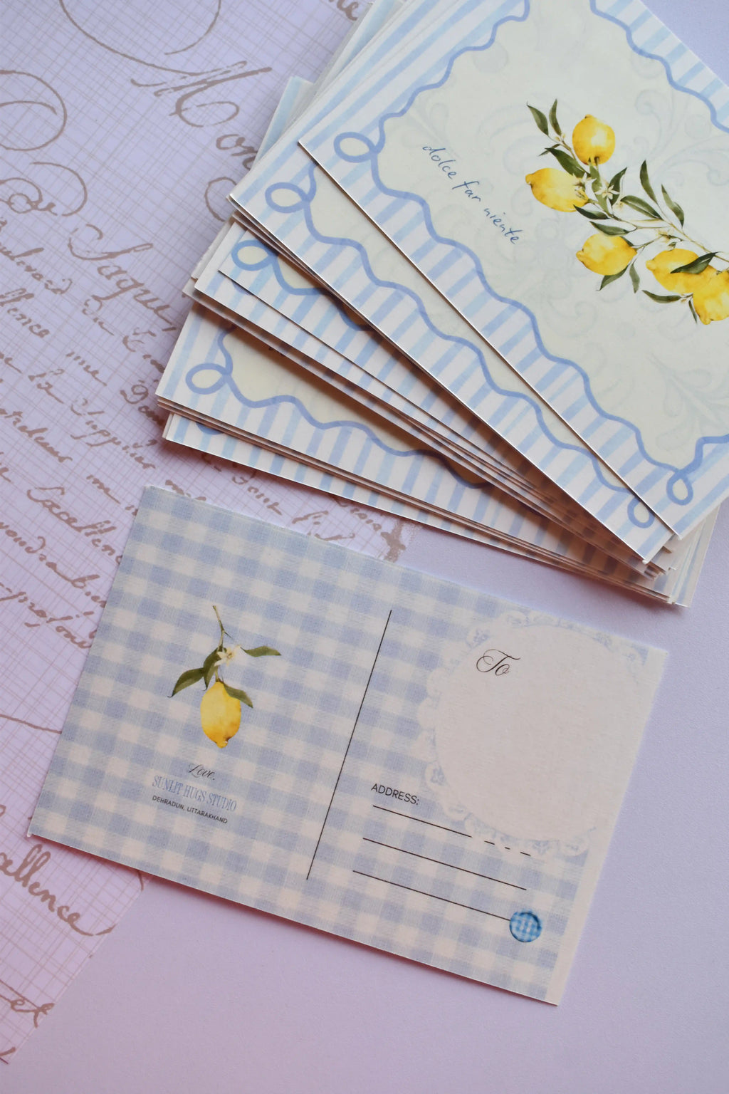 A stack of postcards showing the 'Dolce Far Niente' lemon front design on the top cards. The bottom card is flipped, showing the blue and white wide gingham reverse with the address lines and a single lemon illustration. The stack rests near a purple hand-script paper overlay.