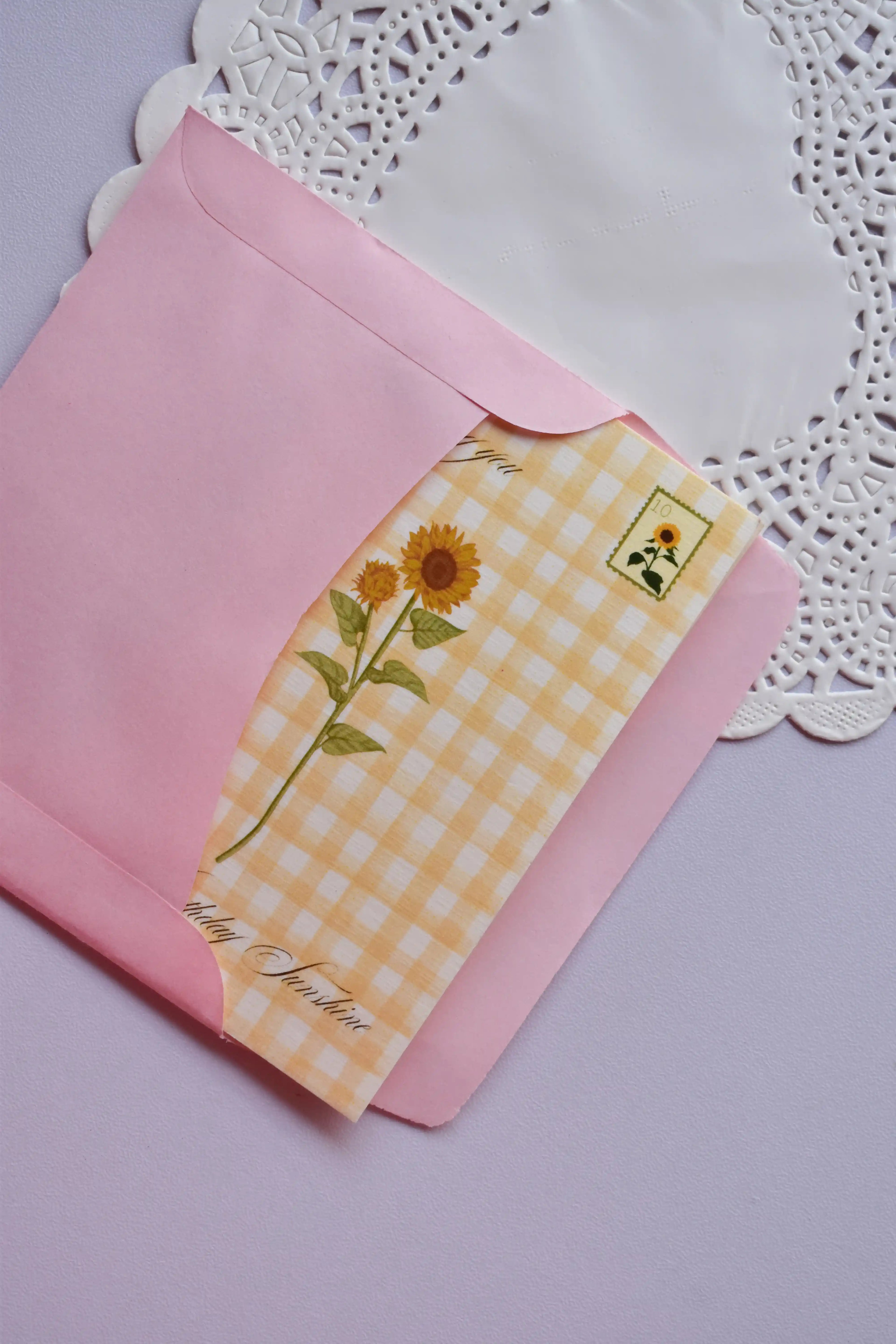 Close-up showing a yellow and white gingham card, decorated with a tall sunflower stem and a tiny sunflower postage stamp design, being gently slipped into a pink envelope. The card edge reads "Wishing you Sunshine