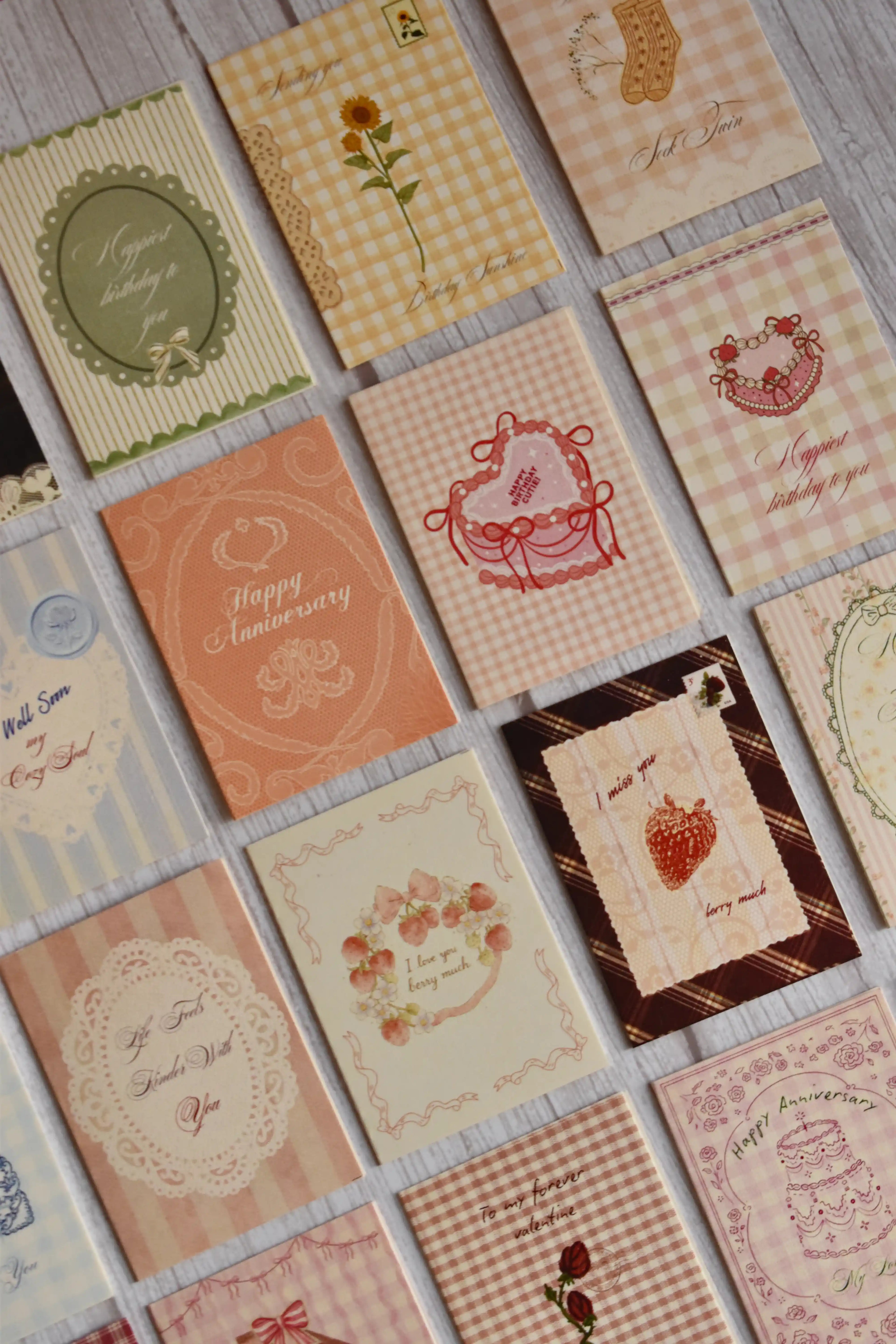 Flat lay of the Sunlit Hugs Assorted Set of 19 Vintage Greeting Cards displayed on a white wooden surface. This cottagecore stationery bundle features gingham, plaid, and floral designs covering multiple occasions including Happy Birthday, Happy Anniversary, Valentine's Day, Get Well Soon, and everyday romantic love notes.