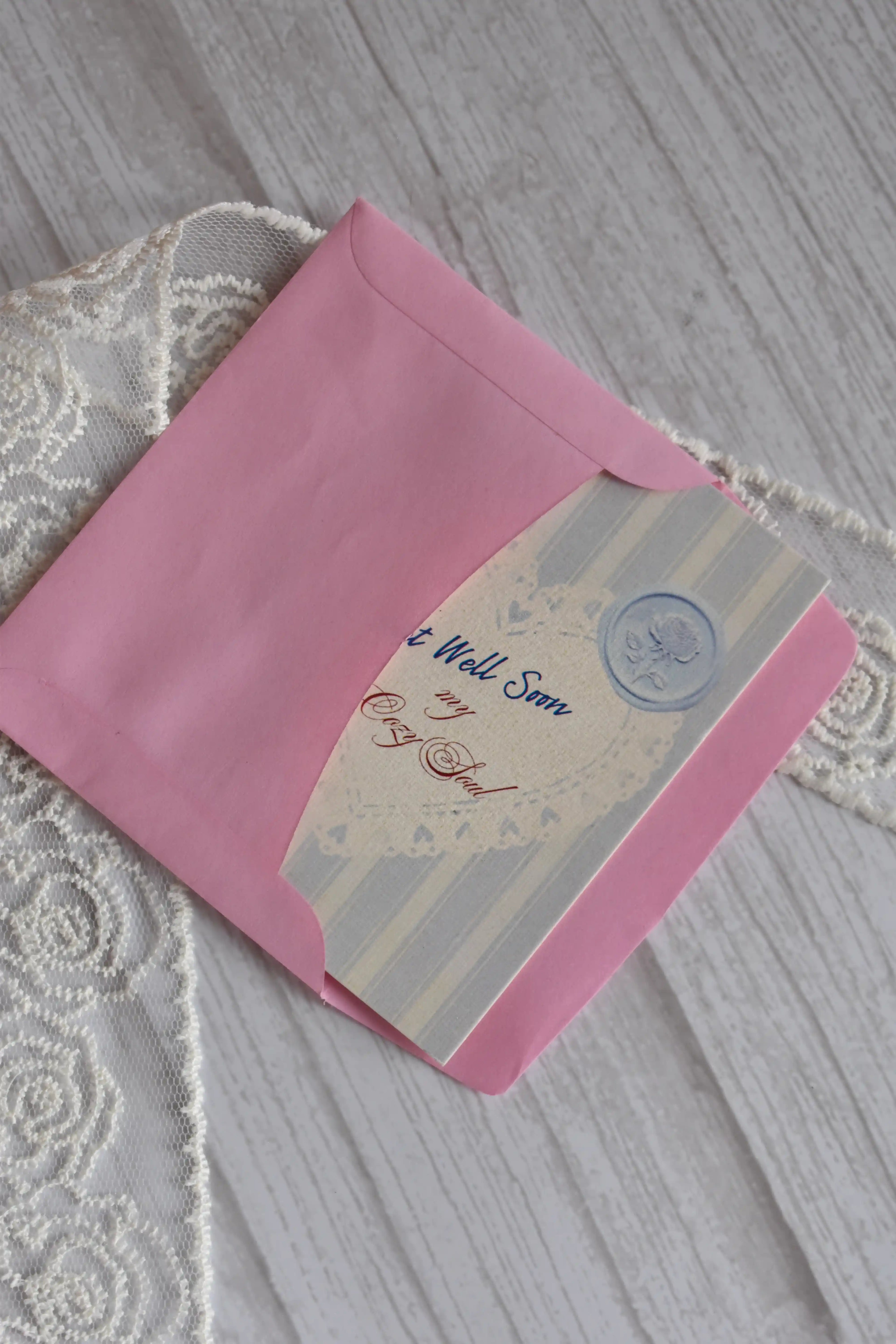Sunlit Hugs Get Well Soon Cozy Soul Greeting Card partially inside a pink envelope on a white wooden surface.
