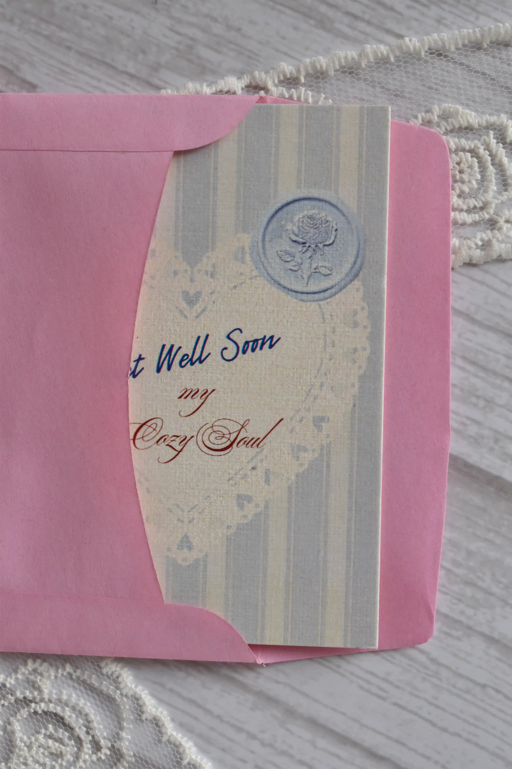 Front cover of the Sunlit Hugs Get Well Soon Cozy Soul Greeting Card resting on a pink envelope, featuring blue stripes, a heart doily design, and "Get Well Soon My Cozy Soul" text.