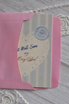 Front cover of the Sunlit Hugs Get Well Soon Cozy Soul Greeting Card resting on a pink envelope, featuring blue stripes, a heart doily design, and "Get Well Soon My Cozy Soul" text.