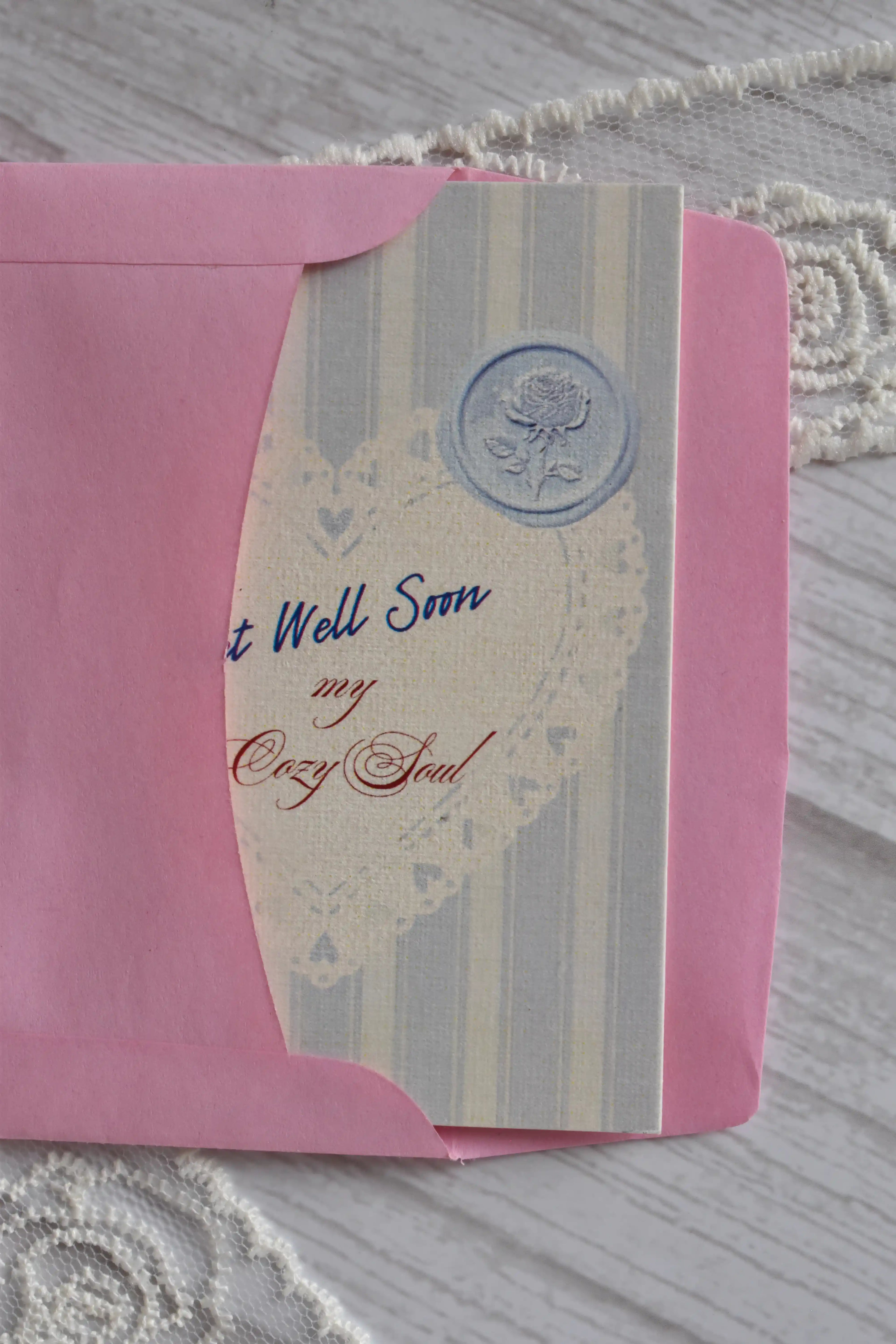 Front cover of the Sunlit Hugs Get Well Soon Cozy Soul Greeting Card resting on a pink envelope, featuring blue stripes, a heart doily design, and "Get Well Soon My Cozy Soul" text.