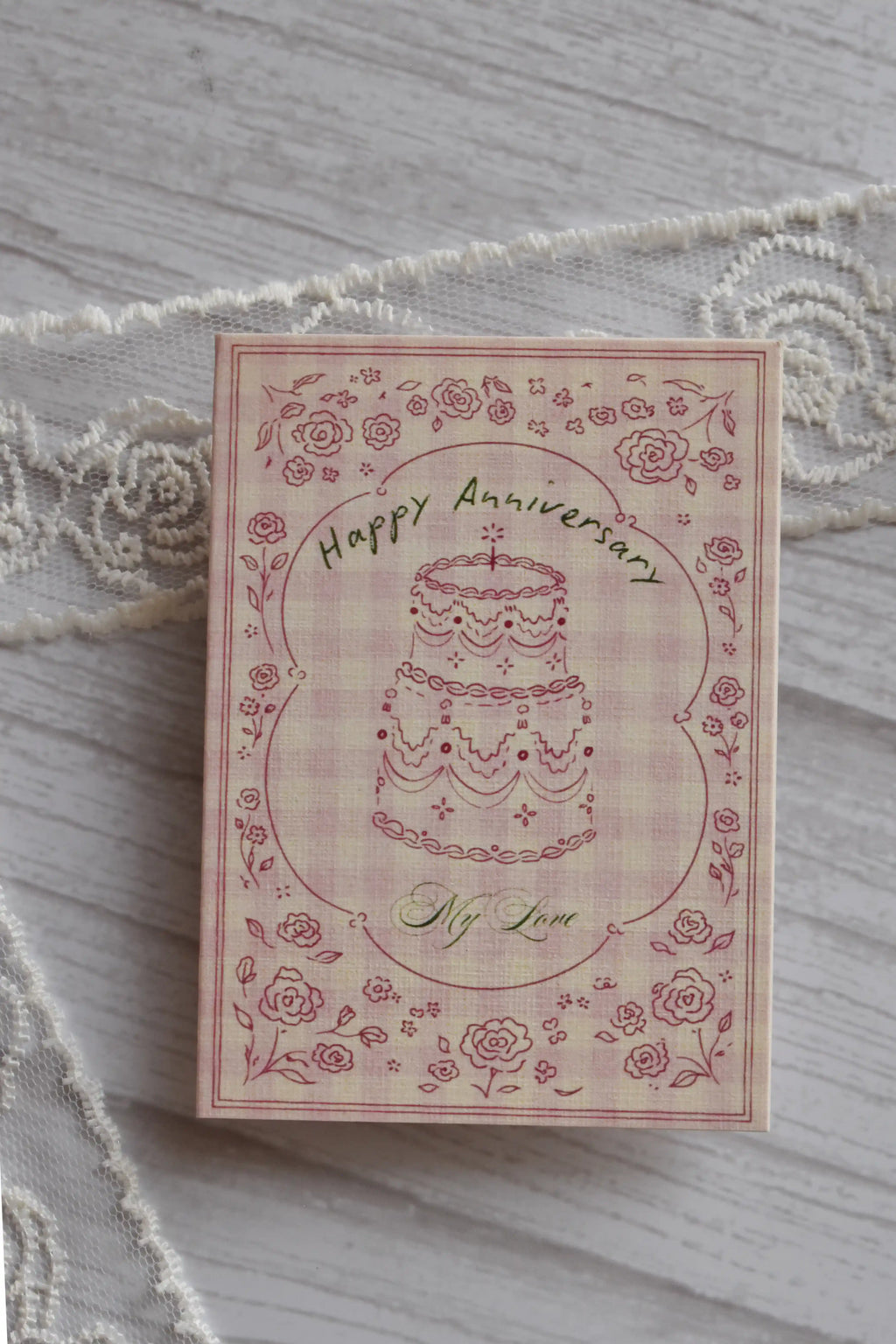 Front cover of the Sunlit Hugs Happy Anniversary Vintage Cake Greeting Card featuring a hand-drawn cake, floral border, and pink gingham background.