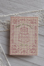 Front cover of the Sunlit Hugs Happy Anniversary Vintage Cake Greeting Card featuring a hand-drawn cake, floral border, and pink gingham background.
