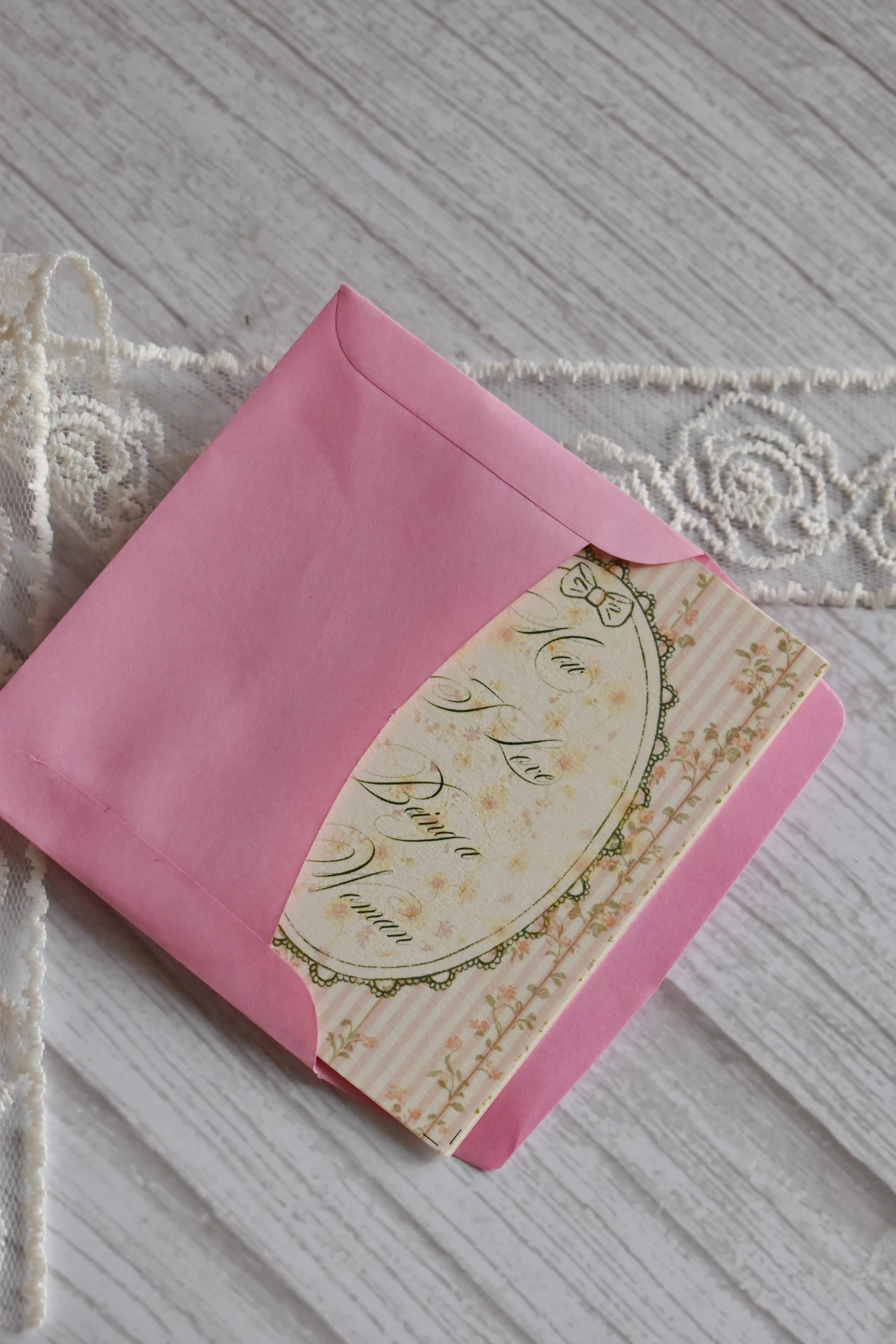Sunlit Hugs How I Love Being a Woman Greeting Card partially tucked inside a bright pink envelope, highlighting the vintage floral frame design.