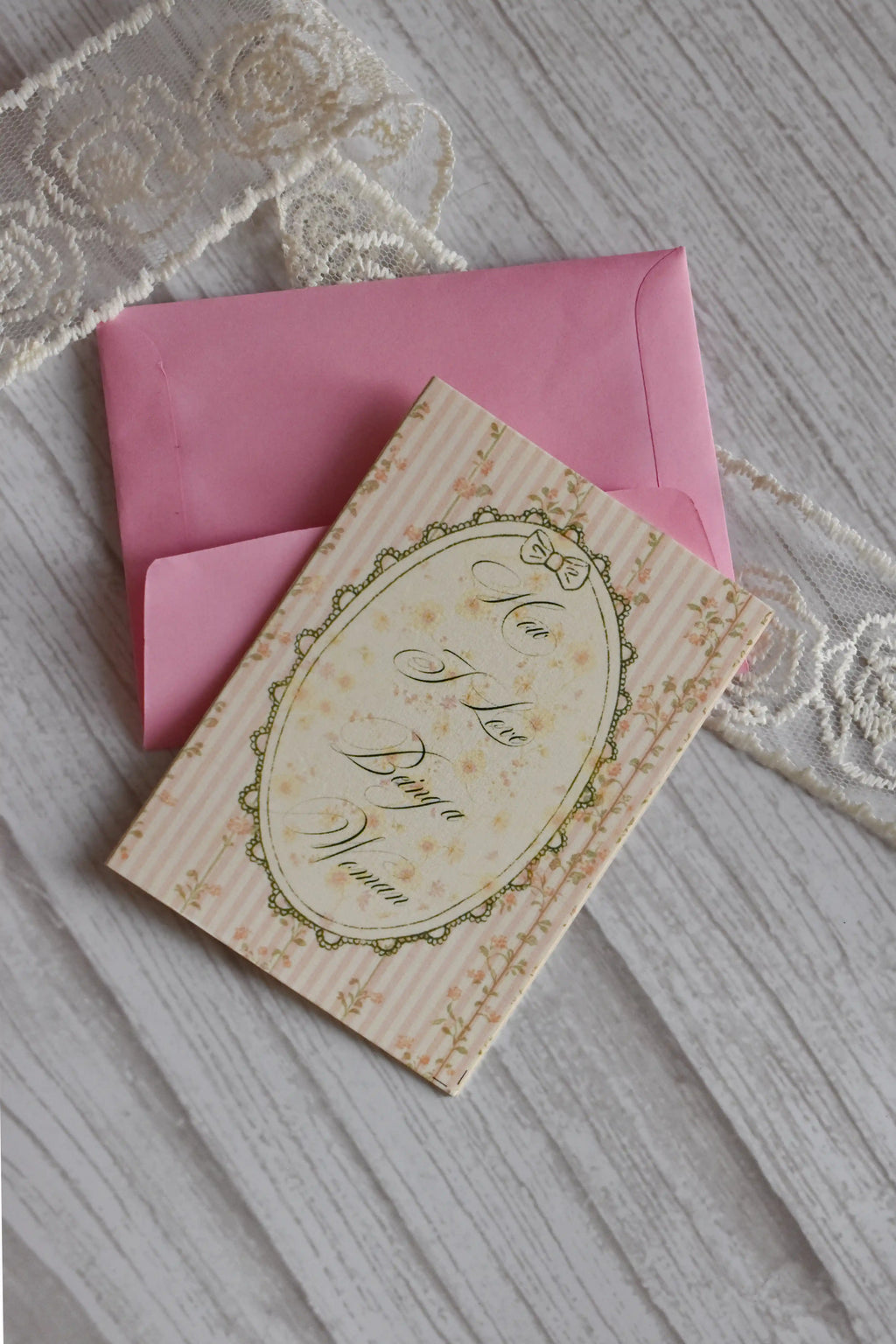 Sunlit Hugs How I Love Being a Woman Greeting Card resting on a matching pink envelope with white floral lace in the background.