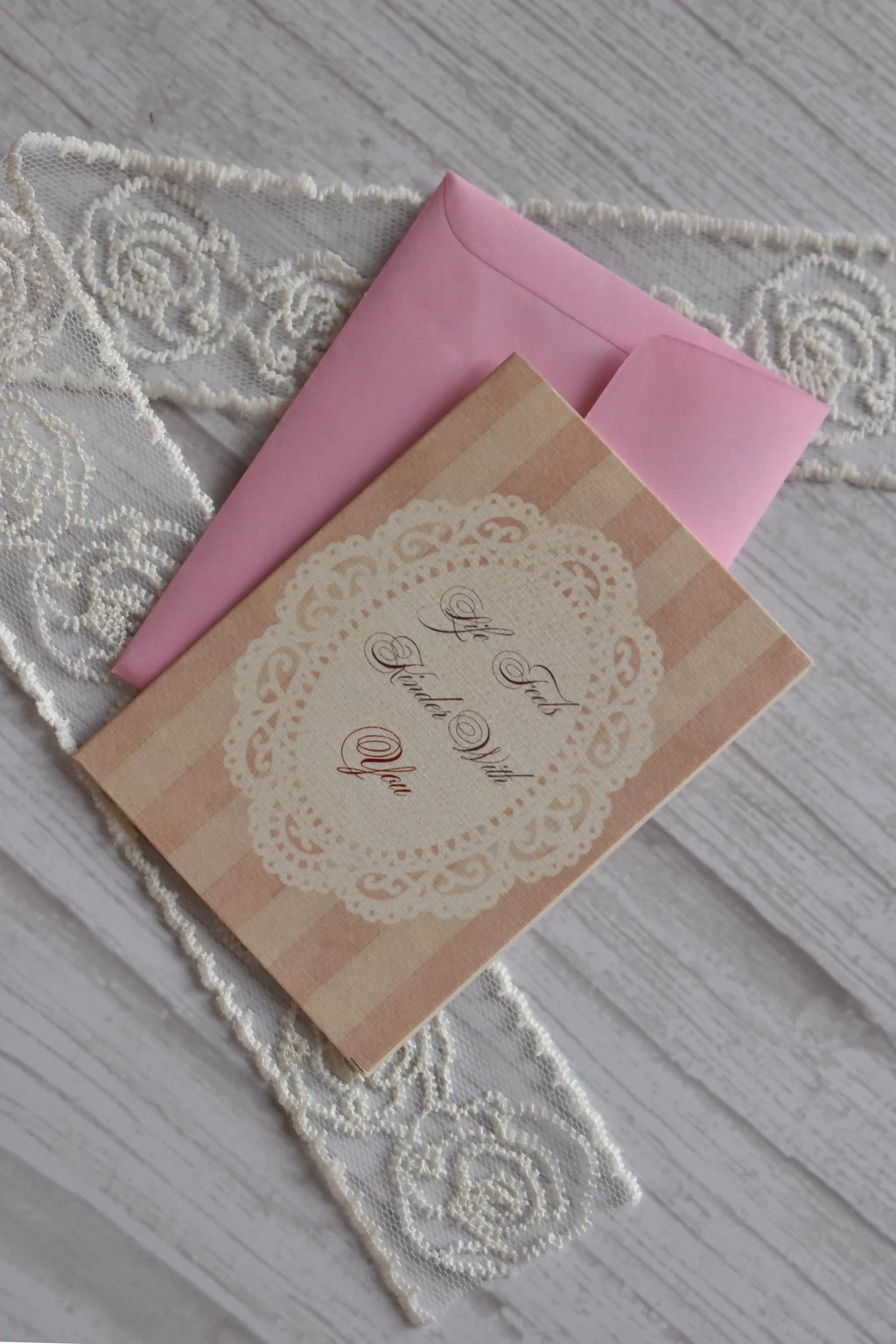 Sunlit Hugs Life Feels Kinder With You Greeting Card resting diagonally on a pink envelope, featuring vintage pink stripes and a white lace doily design next to decorative lace.