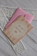 Sunlit Hugs Life Feels Kinder With You Greeting Card resting diagonally on a pink envelope, featuring vintage pink stripes and a white lace doily design next to decorative lace.