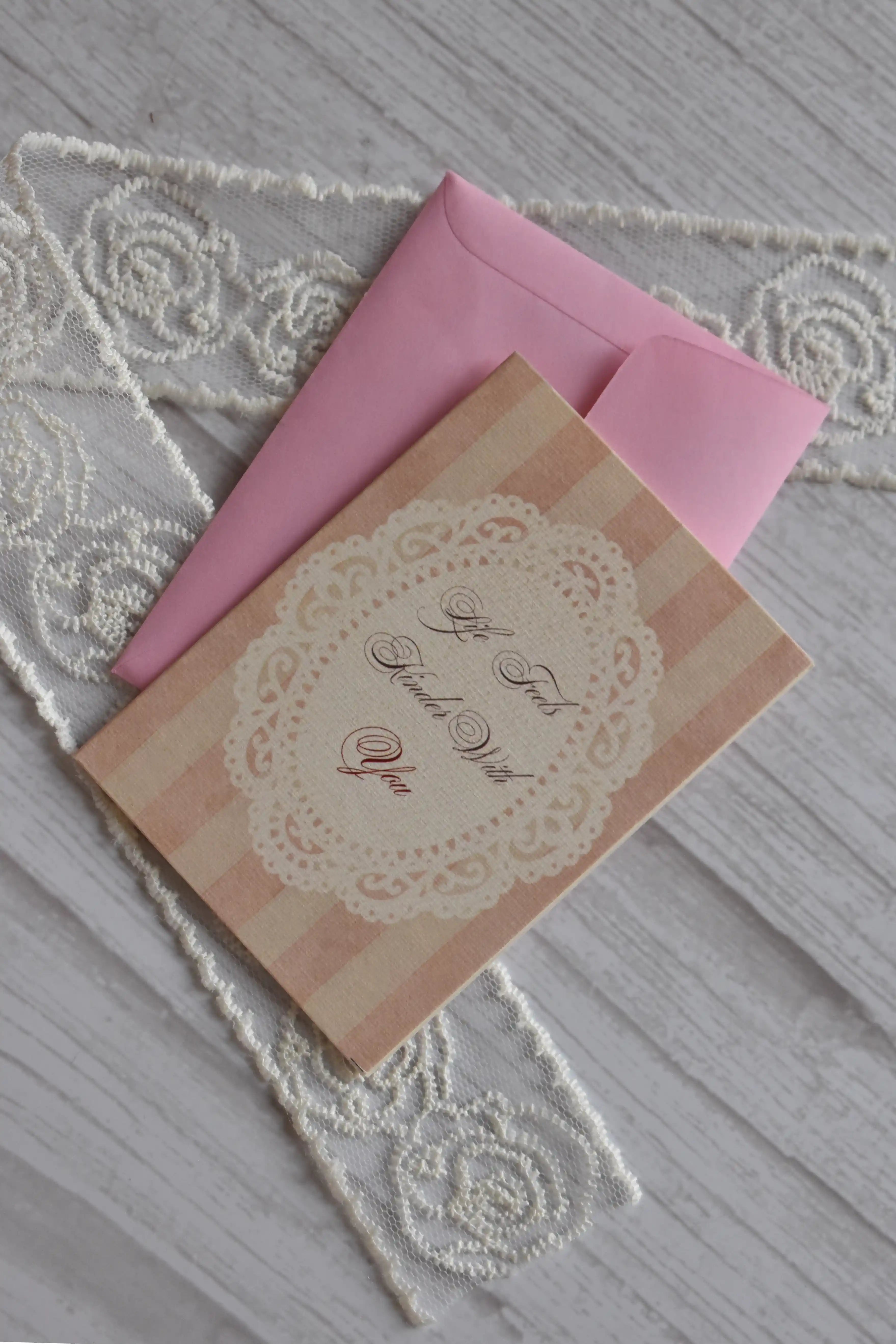 Sunlit Hugs Life Feels Kinder With You Greeting Card resting diagonally on a pink envelope, featuring vintage pink stripes and a white lace doily design next to decorative lace.