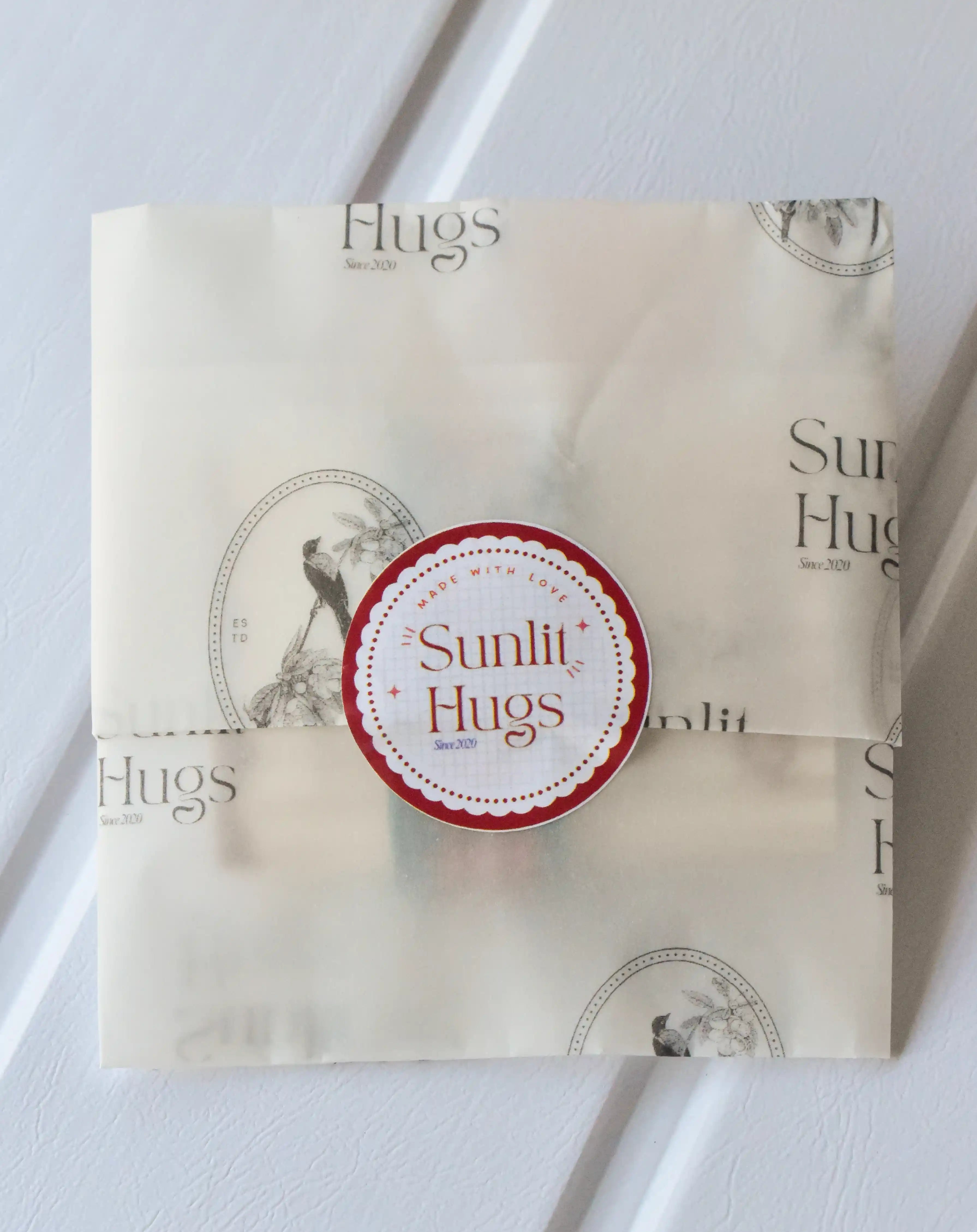 An image of a flat, transparent package of a ribbon, sealed with a circular sticker that reads "Made with Love Sunlit Hugs." It shows an example of the branded packaging.