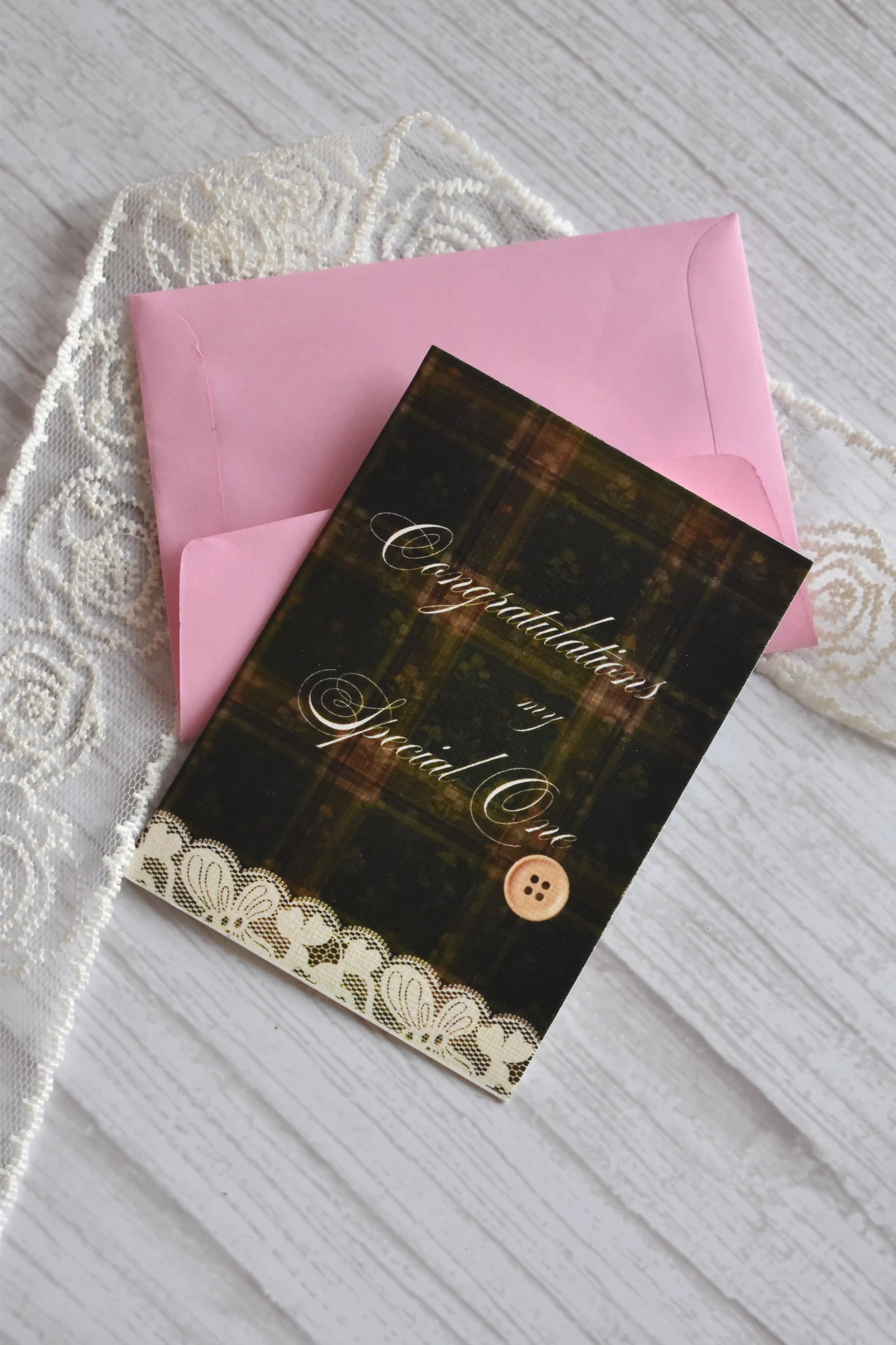 Angled flat lay of the Sunlit Hugs Vintage Plaid Congratulations Greeting Card resting on a pink envelope beside vintage white floral lace.