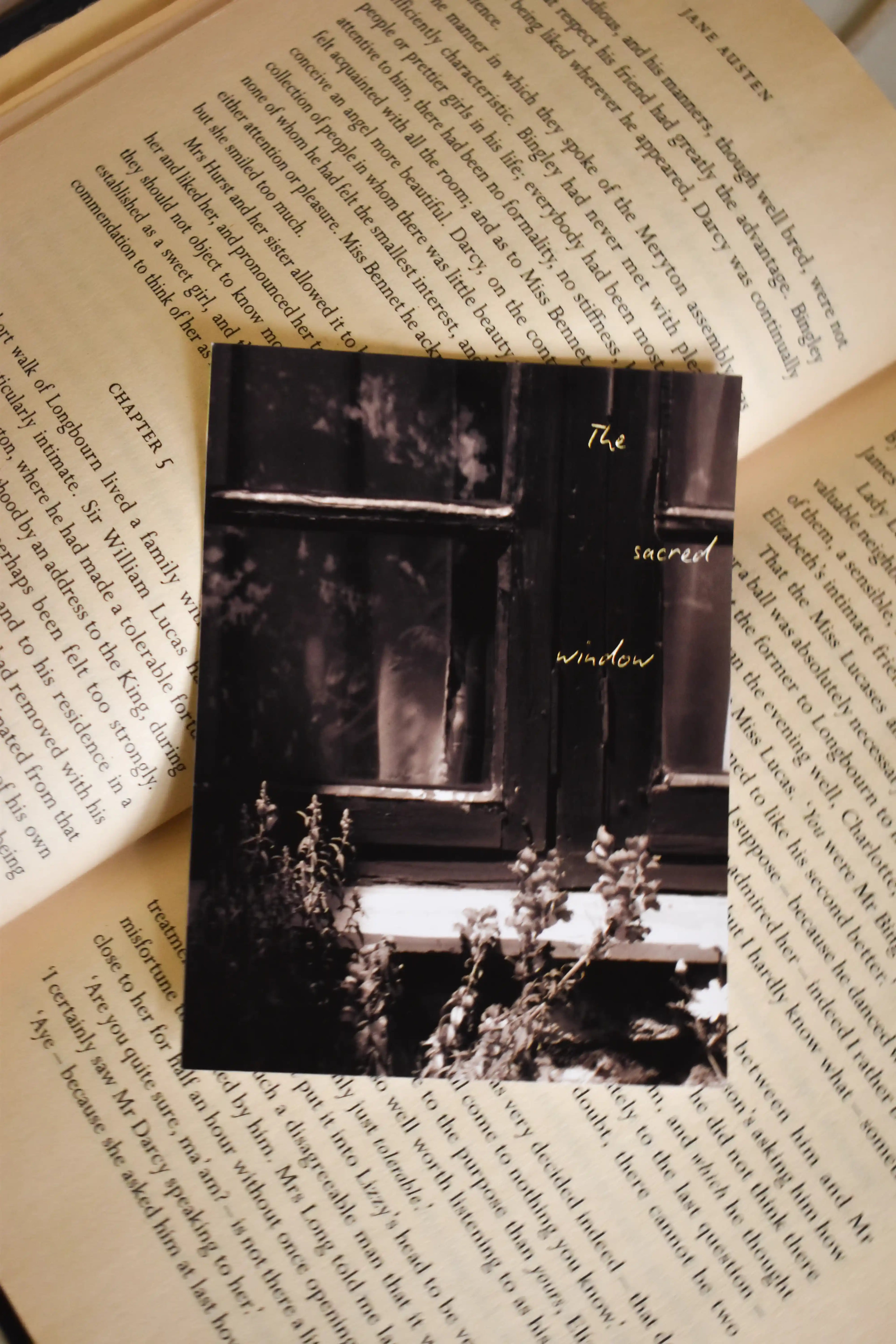Top-down view of the 3x4 inch 'The Sacred Window' Landour Photo Print resting on an open vintage book, showing a moody monochrome window sill with plants and a golden text overlay.