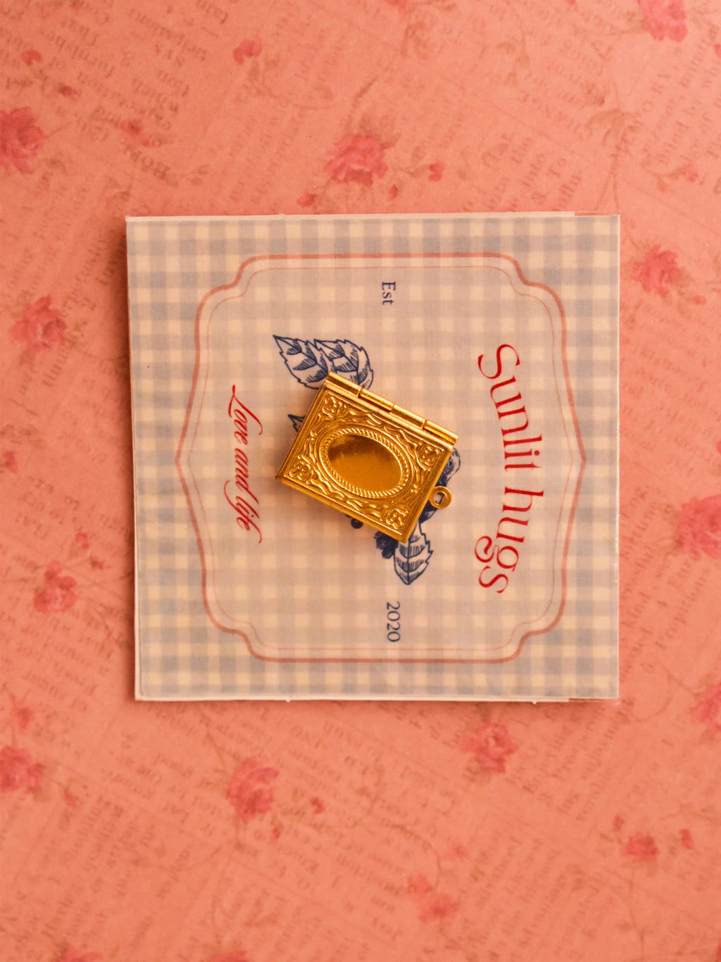 A vintage-style golden book locket placed on a soft blue gingham card with “Sunlit Hugs” branding, styled against a romantic pink floral background.