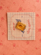 A vintage-style golden book locket placed on a soft blue gingham card with “Sunlit Hugs” branding, styled against a romantic pink floral background.