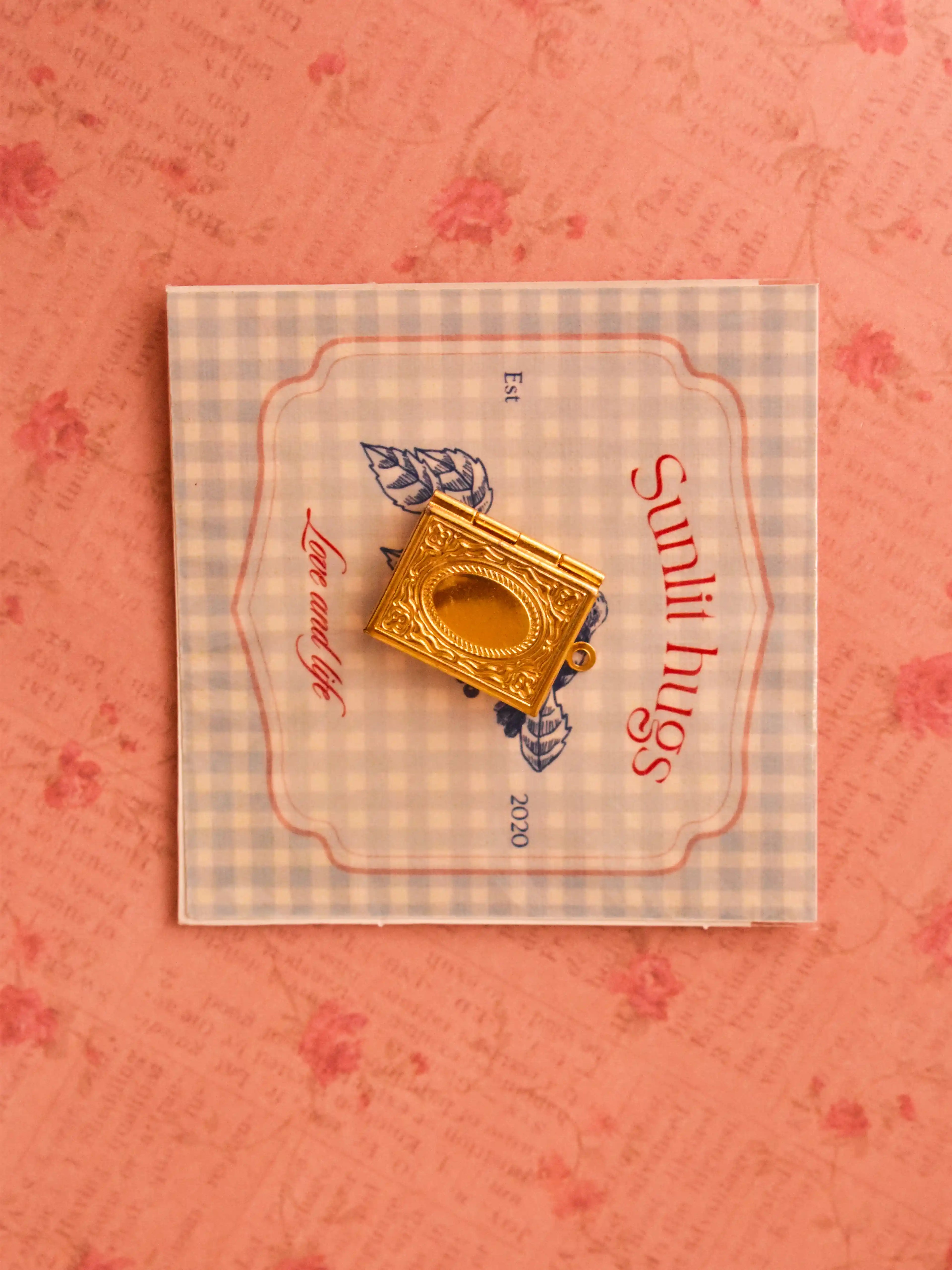A vintage-style golden book locket placed on a soft blue gingham card with “Sunlit Hugs” branding, styled against a romantic pink floral background.