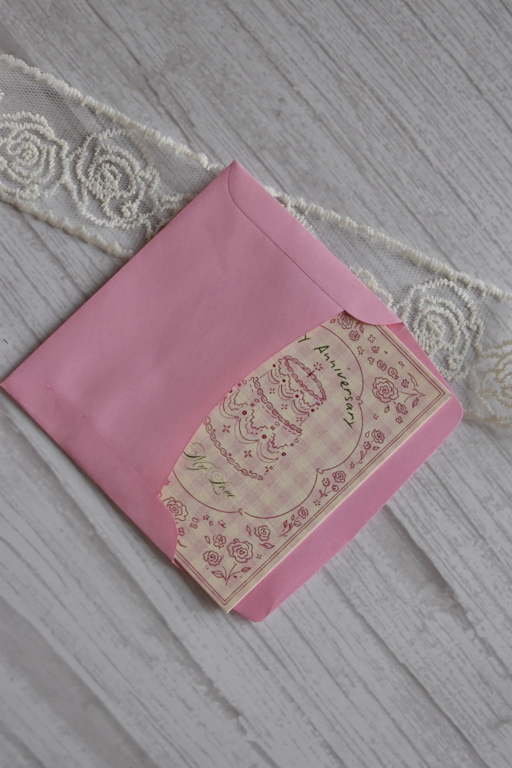 Sunlit Hugs Happy Anniversary Vintage Cake Greeting Card partially tucked inside a bright pink envelope on a white wooden surface.