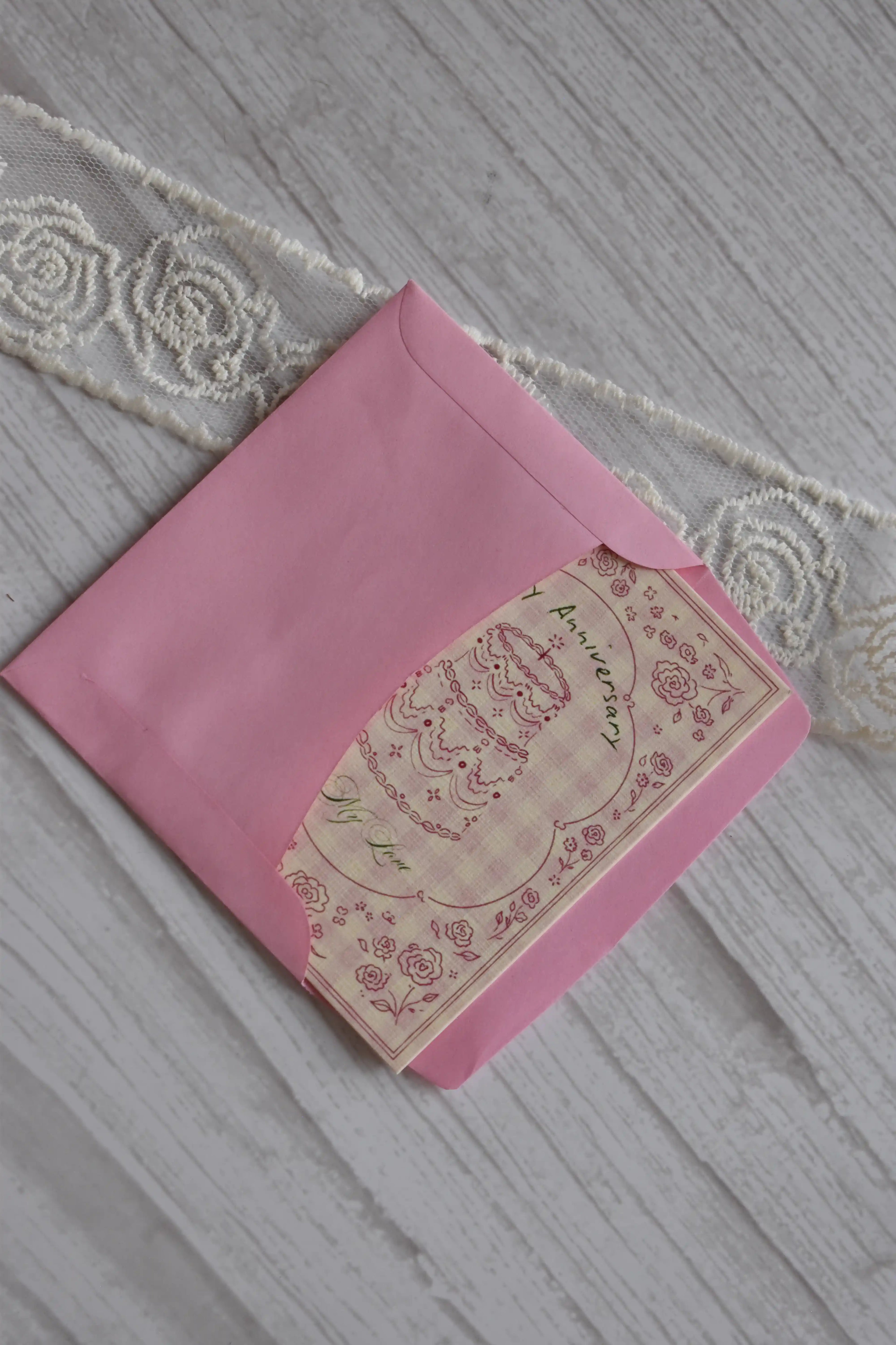 Sunlit Hugs Happy Anniversary Vintage Cake Greeting Card partially tucked inside a bright pink envelope on a white wooden surface.
