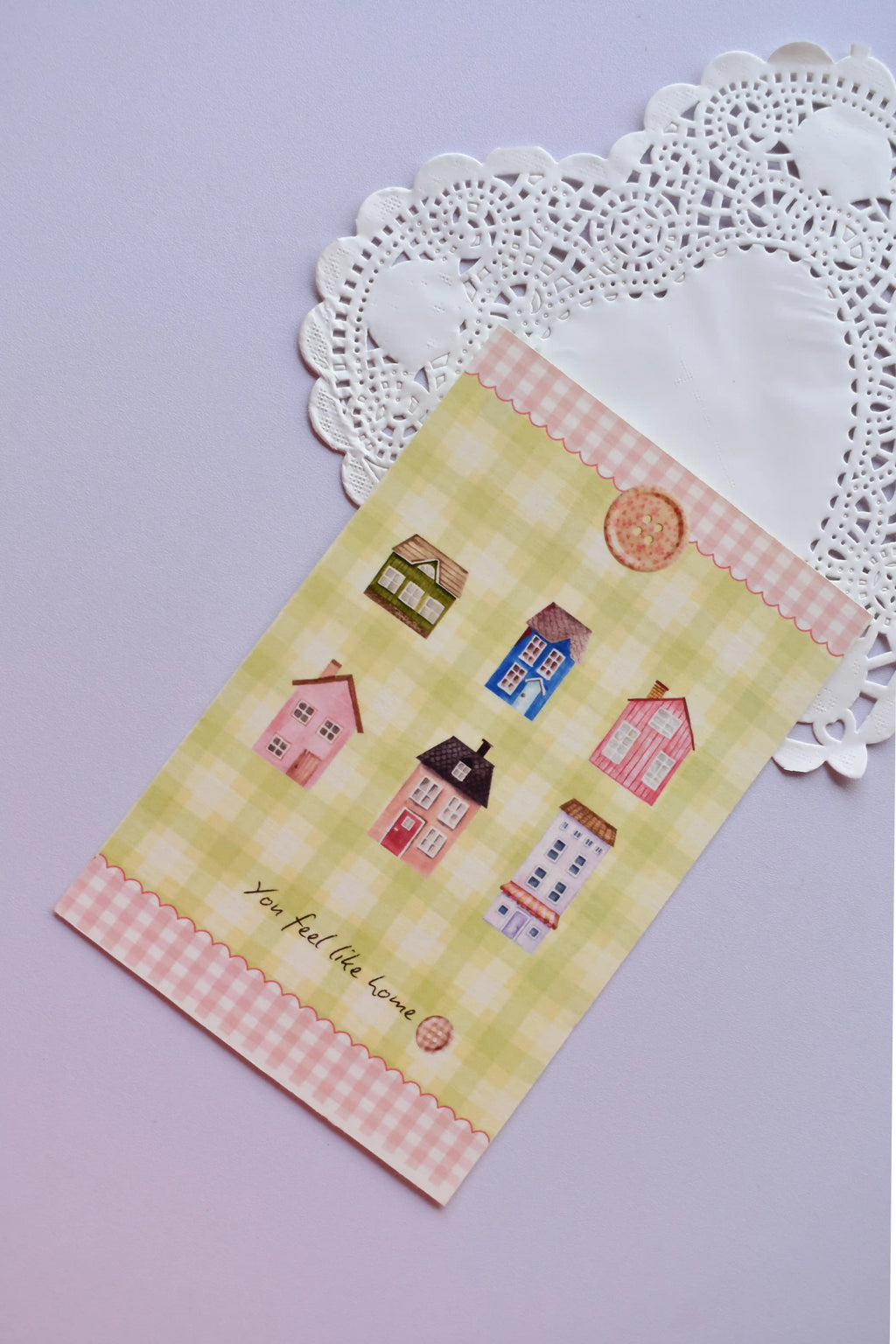 Close-up of a vertical postcard with a pale yellow and green gingham background. It is decorated with six watercolor illustrations of colorful miniature cottages and houses, and features a pink gingham scalloped border. Text at the bottom reads, "You feel like home."