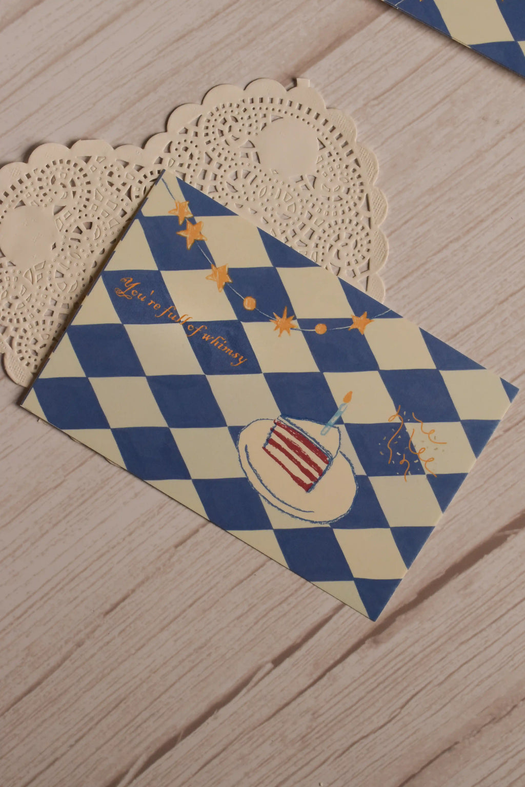 Close view of the postcard front featuring a blue and cream diamond pattern, illustrated birthday cake, candle, star garland and the words You’re Full of Whimsy.