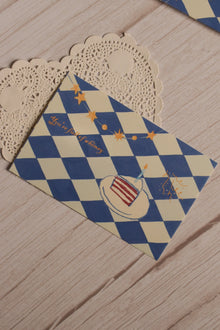Close view of the postcard front featuring a blue and cream diamond pattern, illustrated birthday cake, candle, star garland and the words You’re Full of Whimsy.