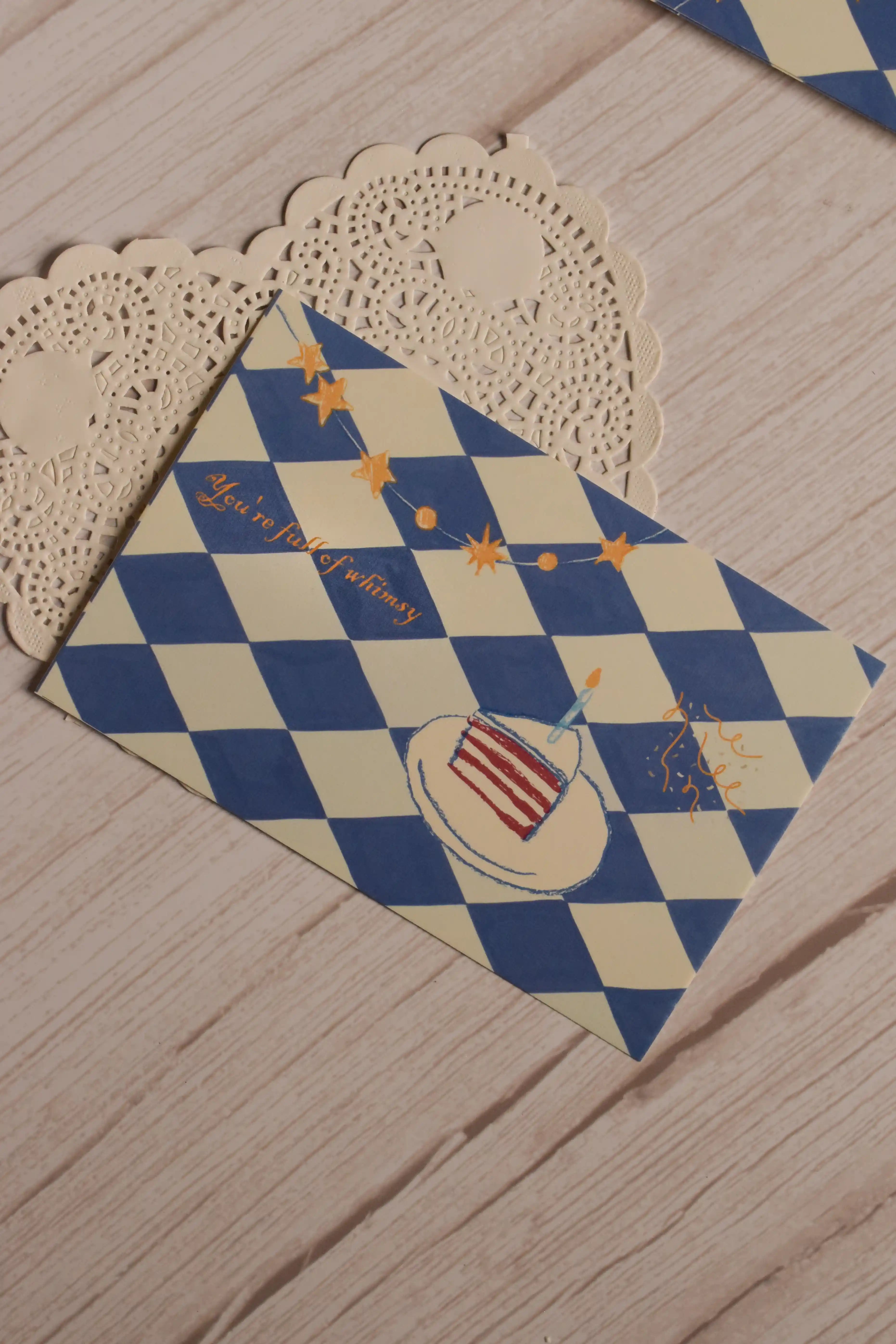 Close view of the postcard front featuring a blue and cream diamond pattern, illustrated birthday cake, candle, star garland and the words You’re Full of Whimsy.