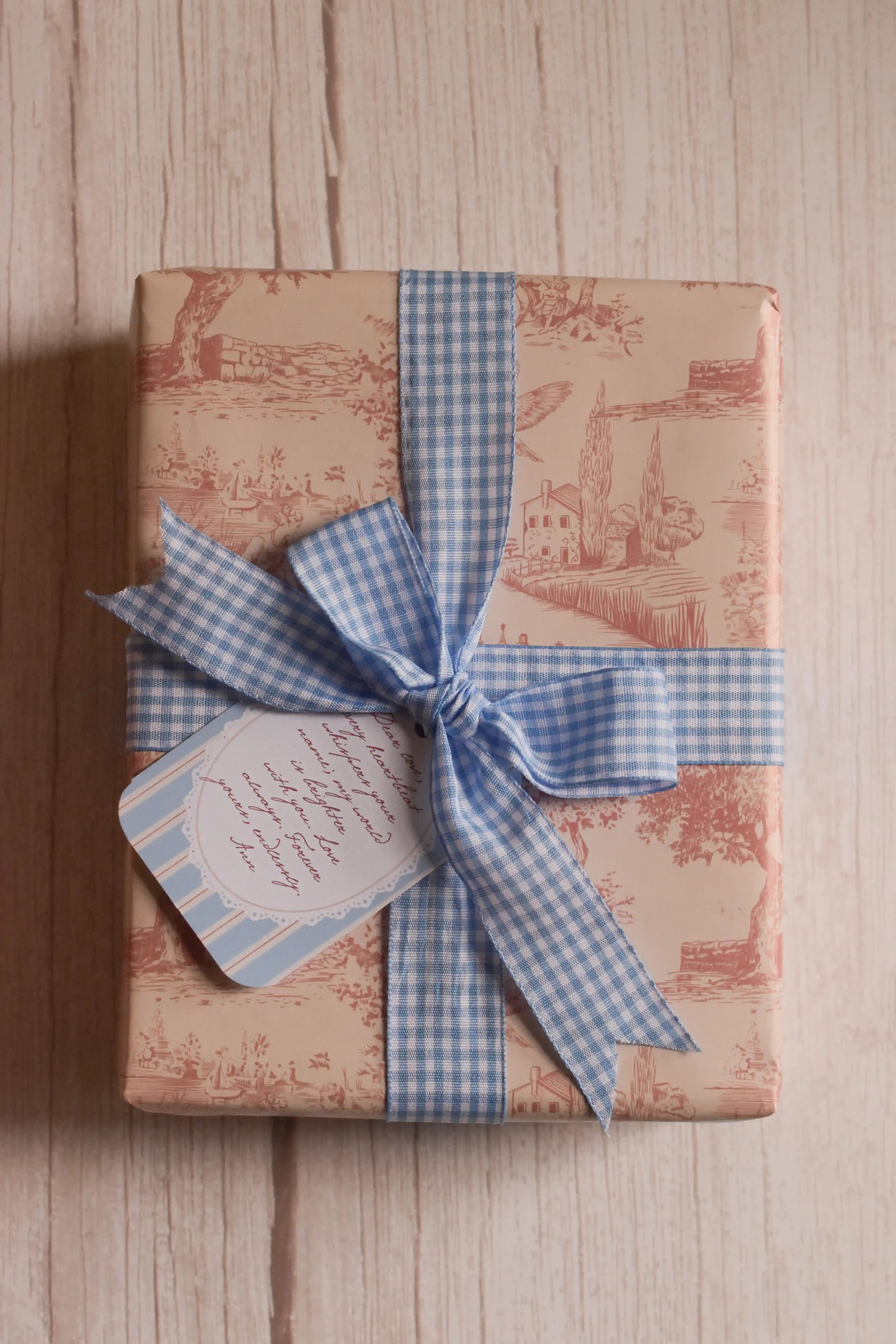 Top view of a rectangular gift wrapped in vintage toile paper featuring countryside scenes, tied with a wide powder blue gingham ribbon and finished with a coordinating message tag.