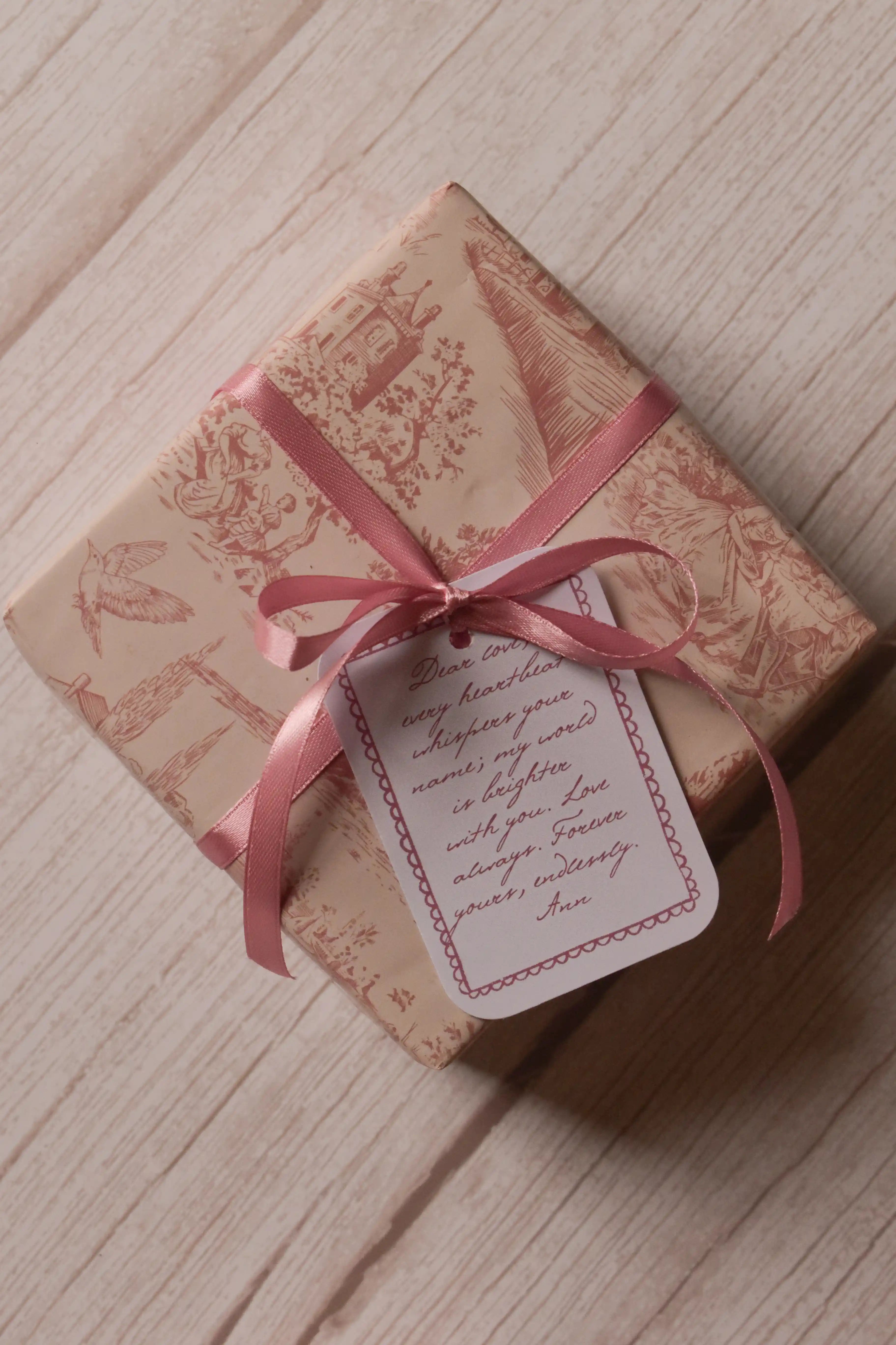 A top view of a rectangular gift wrapped in cream and dusty rose toile paper, finished with a coordinating rose pink ribbon bow and a decorative message tag.