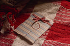 The set of four birthday postcards arranged in its striped paper sleeve with a red gingham ribbon, photographed against a textured red, cream and brown woven fabric background.