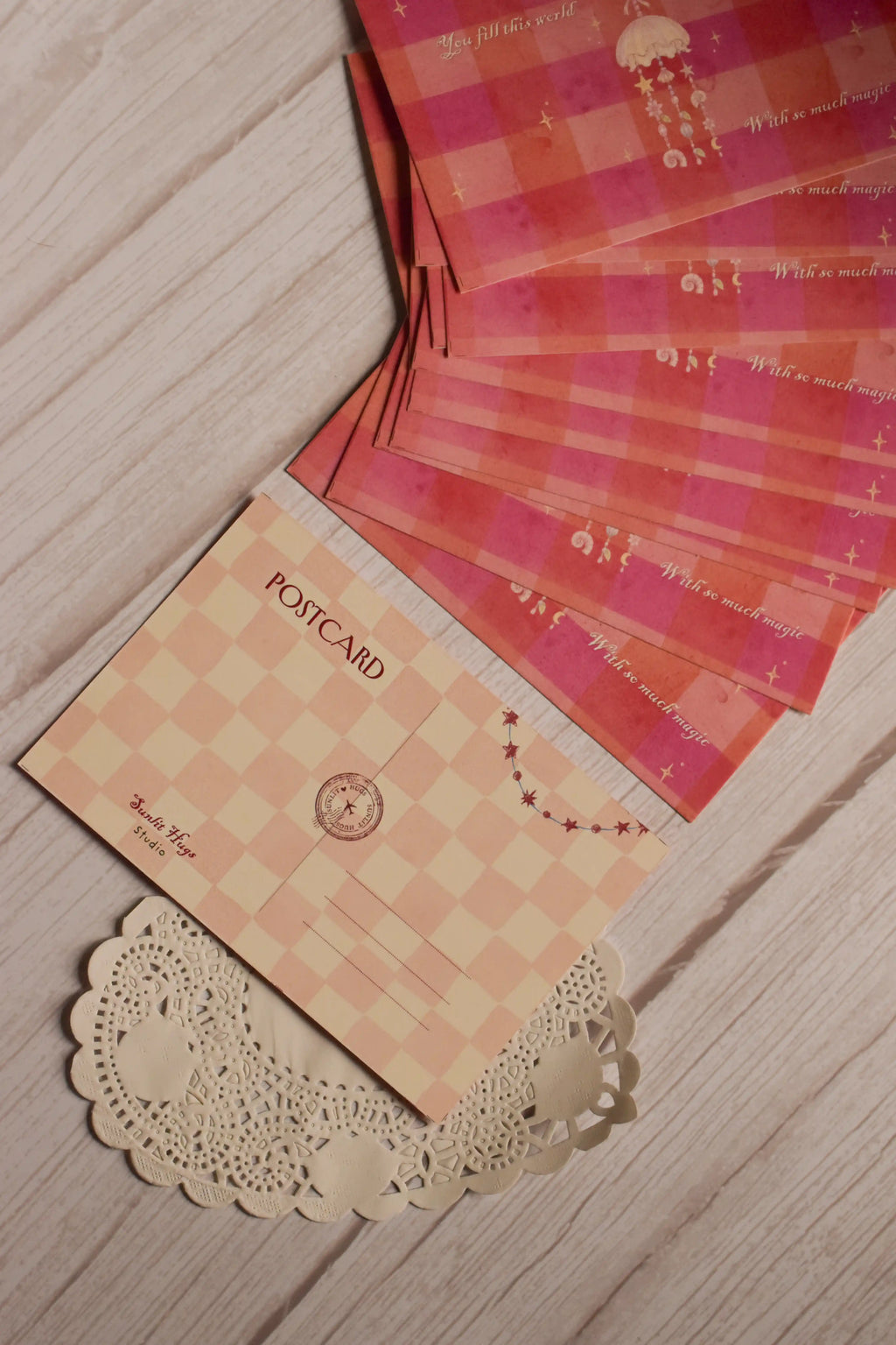 Back view of the 4 x 6 inch postcard featuring a soft blush and cream checkered pattern, POSTCARD heading, address lines, circular postal-style mark, Sunlit Hugs Studio branding and a small star garland.