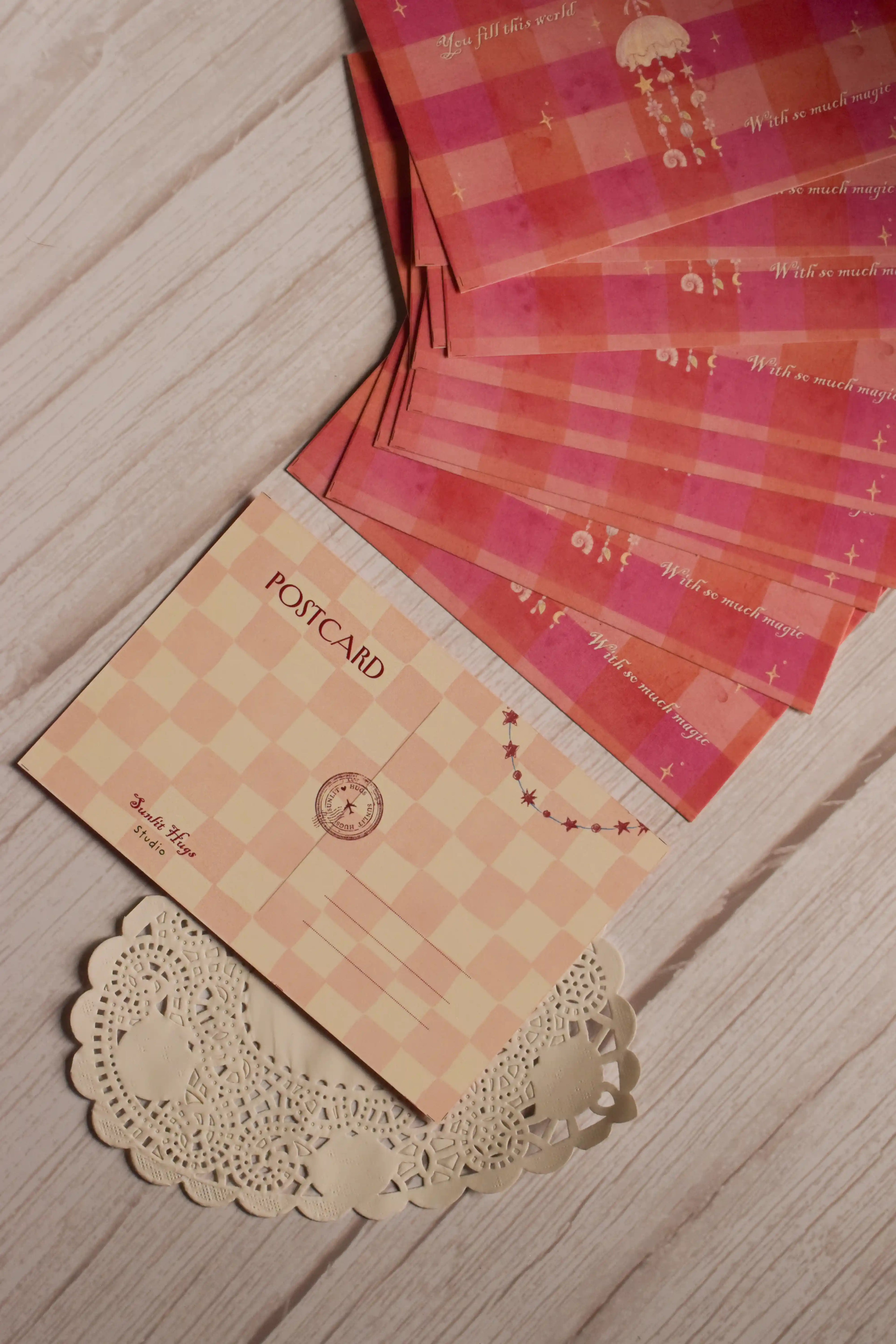 Back view of the 4 x 6 inch postcard featuring a soft blush and cream checkered pattern, POSTCARD heading, address lines, circular postal-style mark, Sunlit Hugs Studio branding and a small star garland.